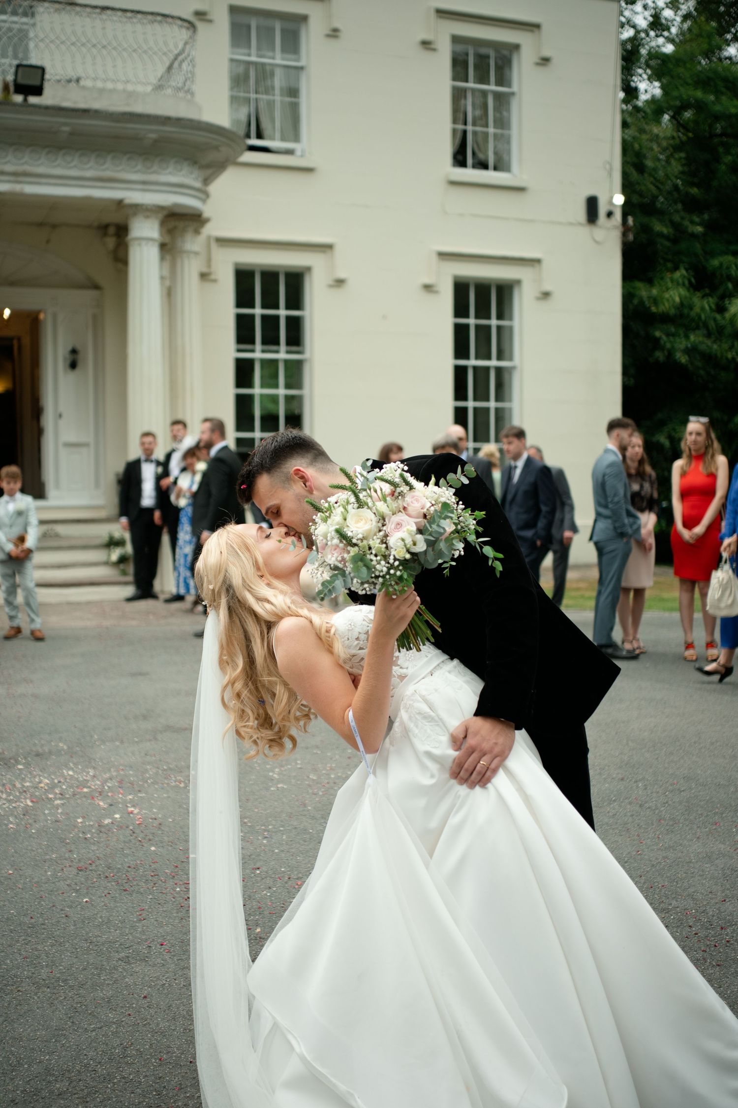 Groom dipping bride for romantic kiss outside elegant white manor wedding venue.