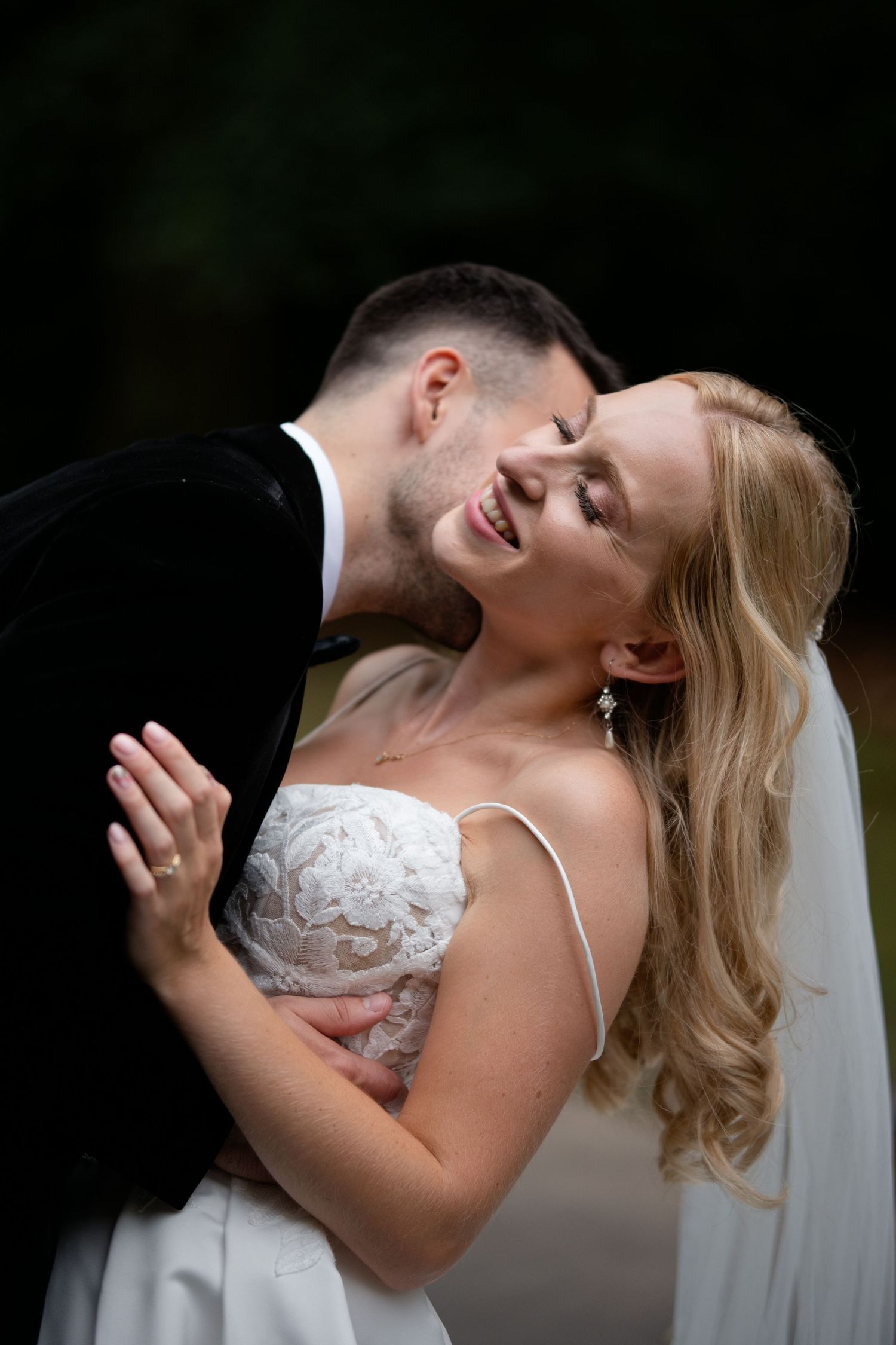 Groom tenderly kisses bride on the cheek as she smiles joyfully in her lace wedding dress outdoors.