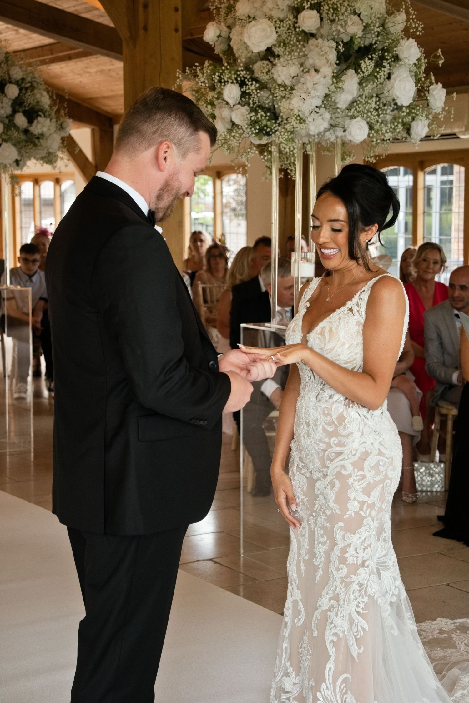 Joyful bride in lace gown places wedding ring on grooms finger during ceremony surrounded by white floral arrangements.