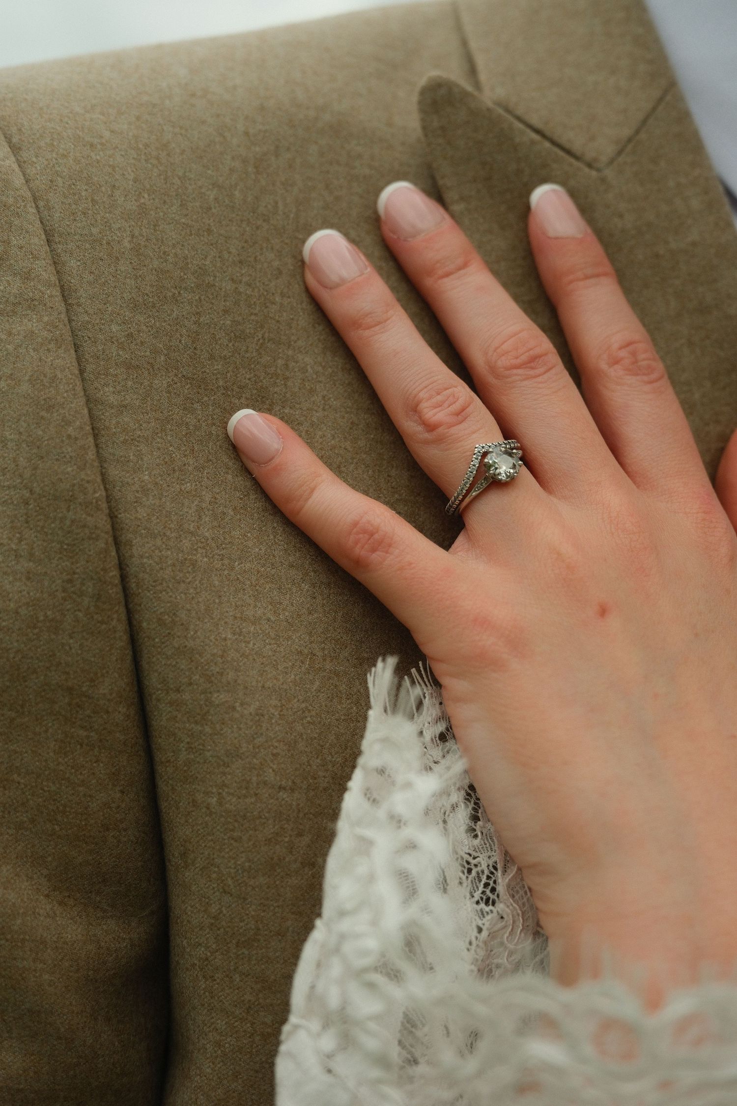 Close-up of bride's hand with diamond engagement ring resting on groom's tan suit jacket.
