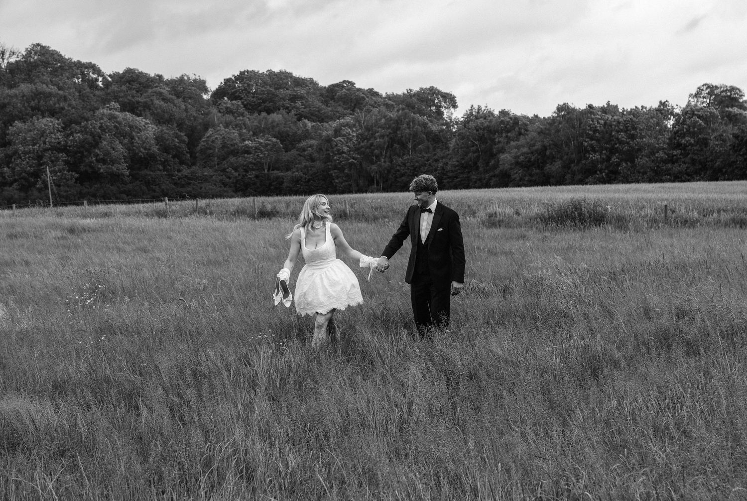 Black and white photo of a bride and groom holding hands walking through a lush meadow with trees in the background.
