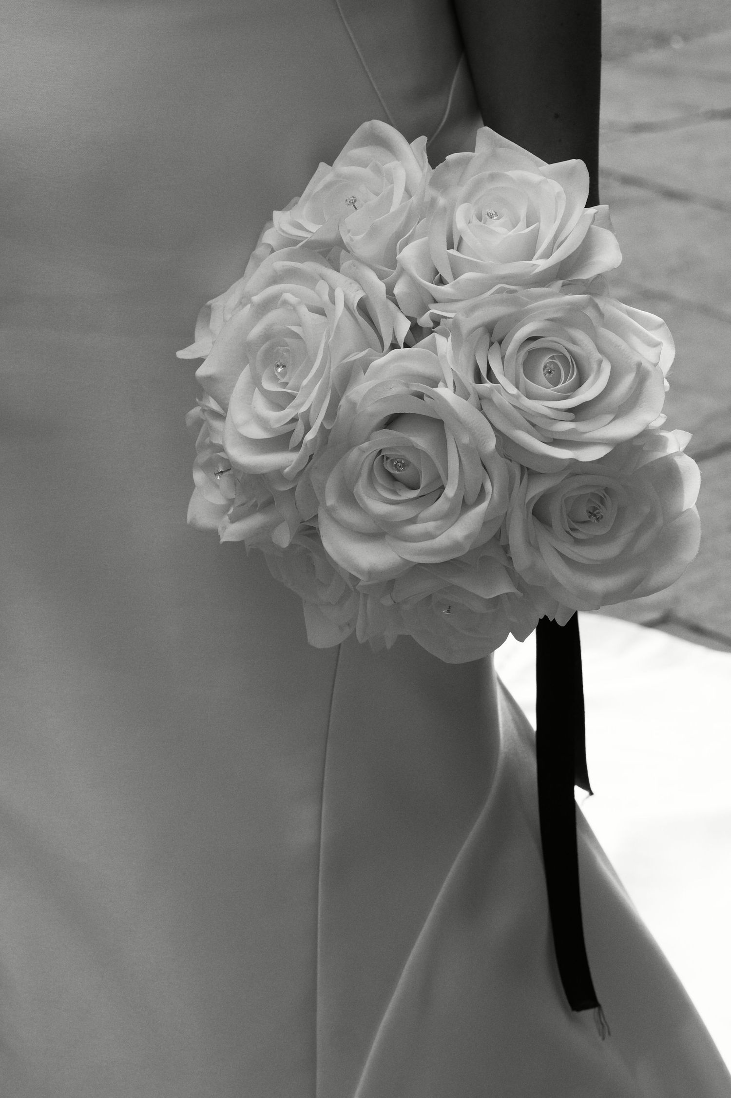 Black and white photo of bride holding elegant white rose bouquet with dark ribbon in wedding dress.