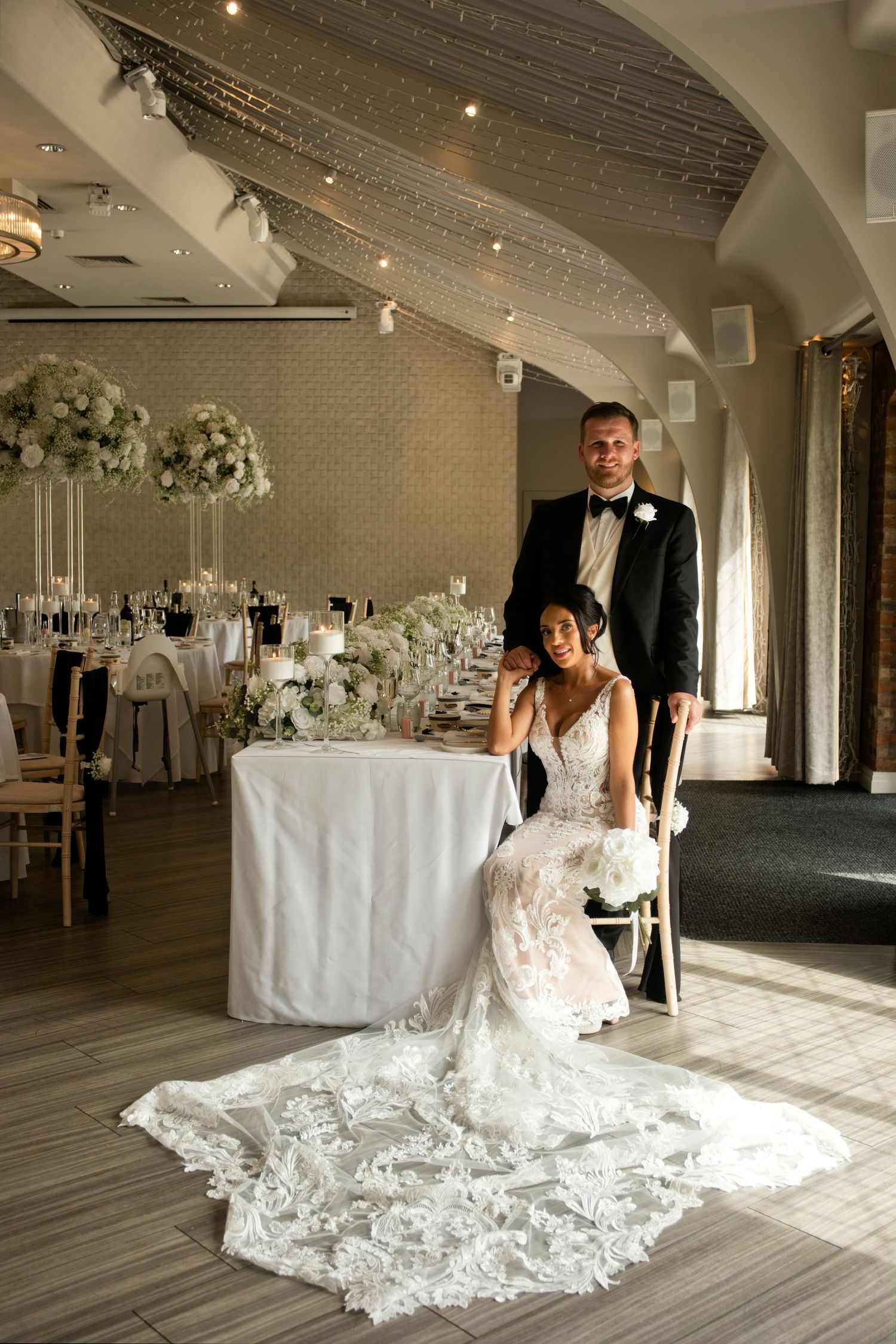 Bride in lace gown seated beside groom in tuxedo at elegant white floral wedding reception.