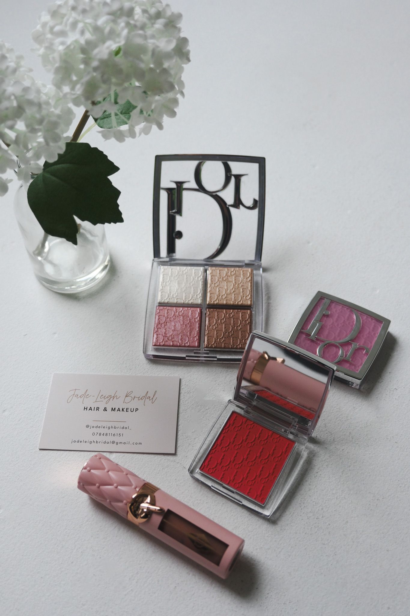 Dior makeup collection featuring eyeshadow palette, blush compacts, and pink lipstick beside a business card and white hydrangeas.