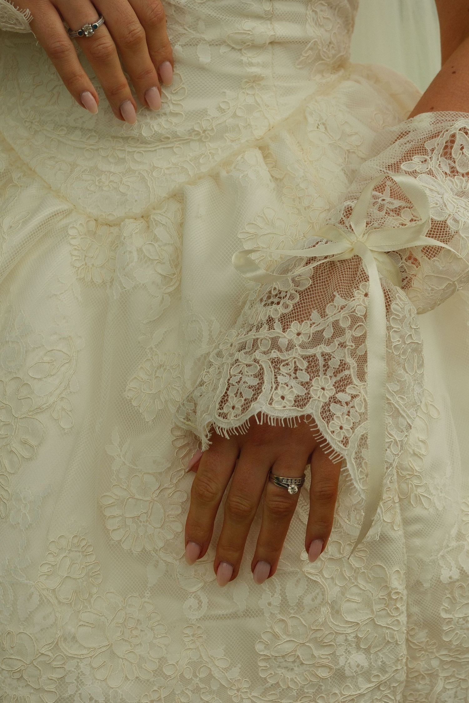 Close-up of bride's hands in lace gloves showing diamond engagement ring on white gown.