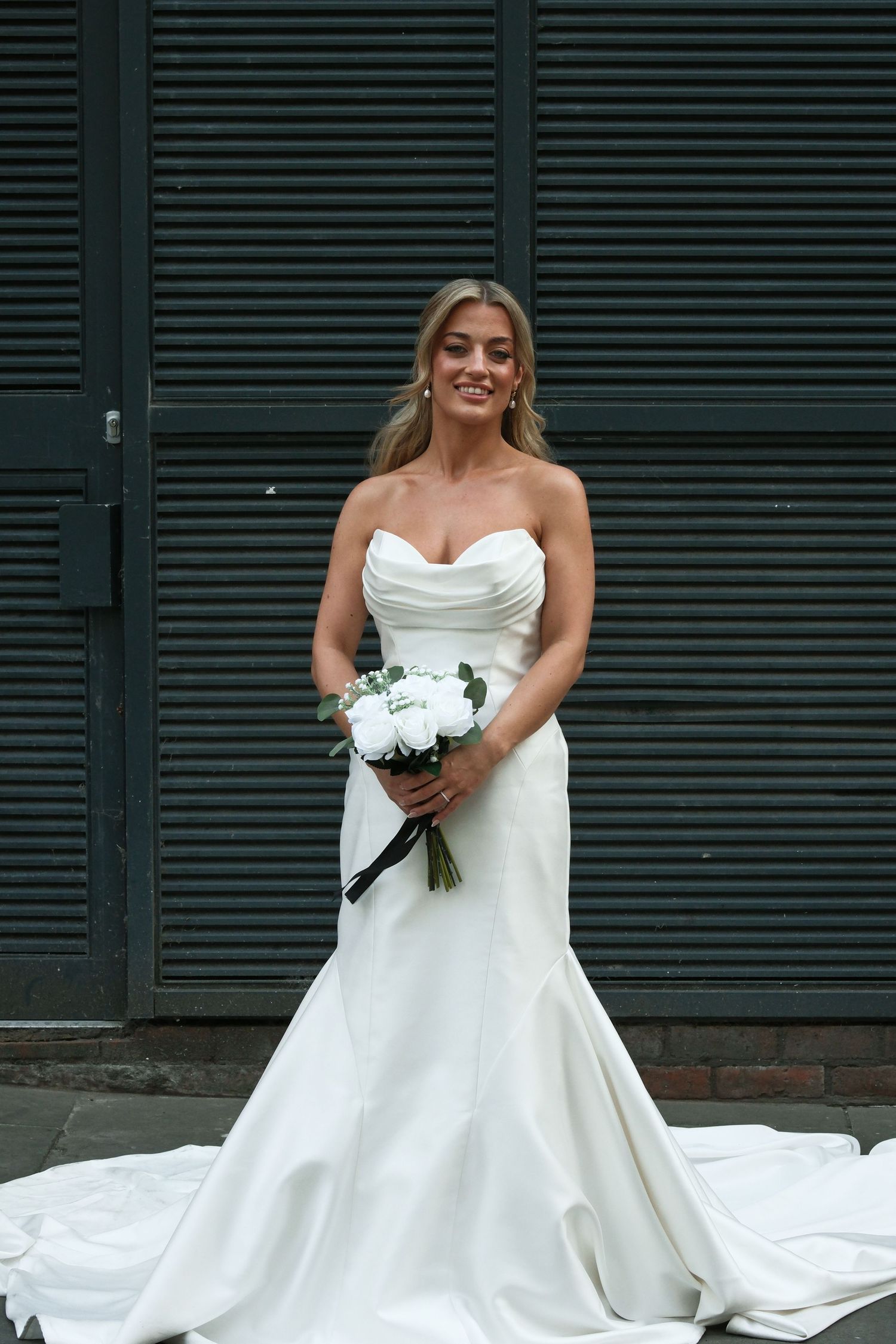 Elegant bride in strapless white mermaid wedding gown holds white rose bouquet against dark shuttered backdrop.