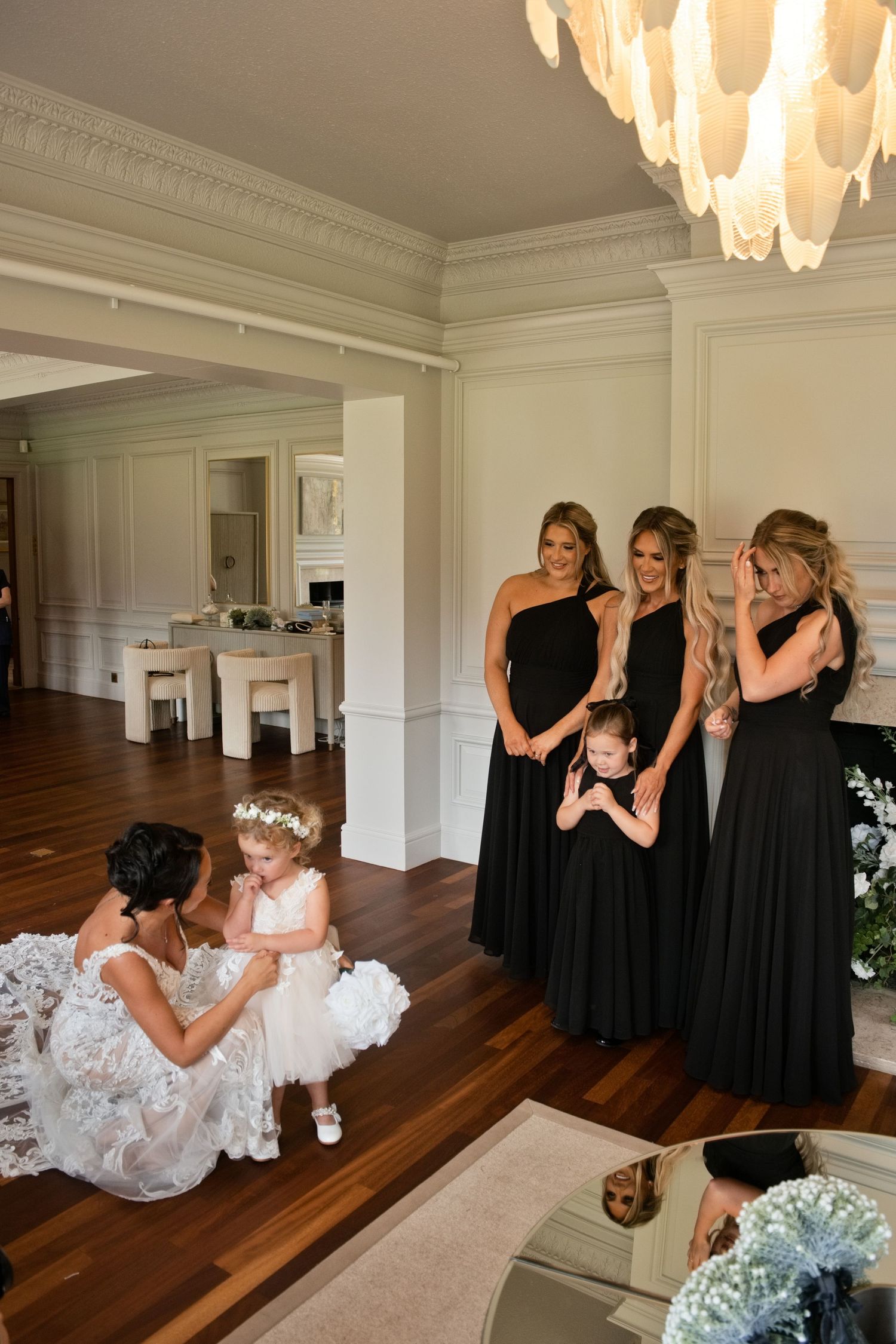 Bride kneeling with flower girl in white dress while three bridesmaids in black gowns smile in elegant venue.