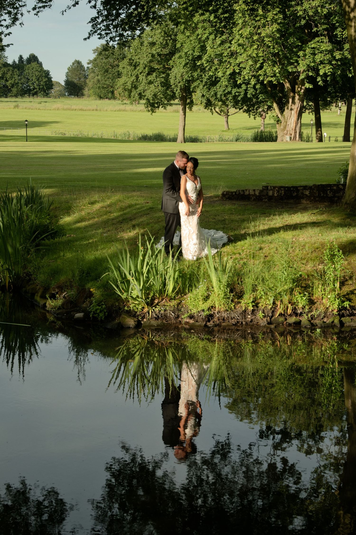 Bride and groom embracing by pond with their reflection visible in water.