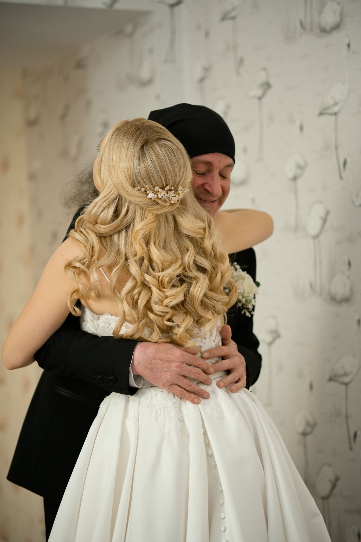 Emotional father hugging bride in white wedding dress with elegant half-up curly blonde hairstyle.