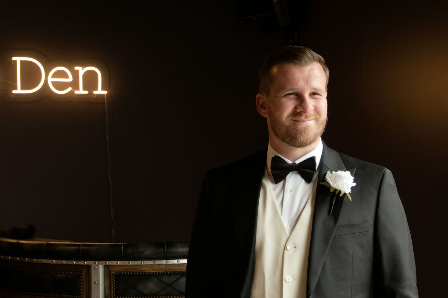 Handsome groom in black tuxedo with white boutonniere smiling beside glowing Den neon sign.