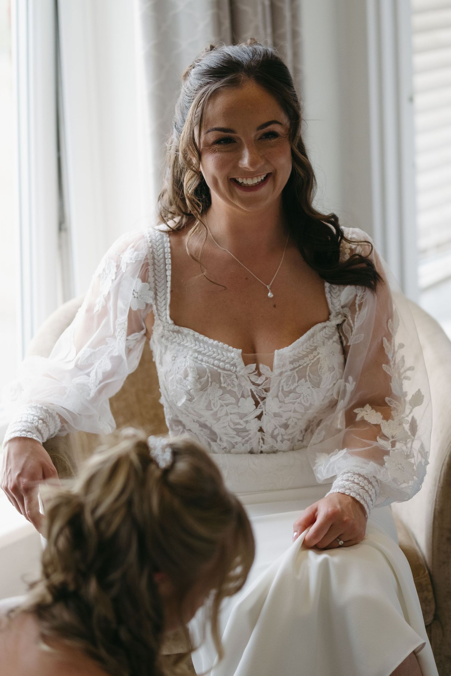 Smiling bride in lace long-sleeve wedding dress seated by bright window on wedding day.