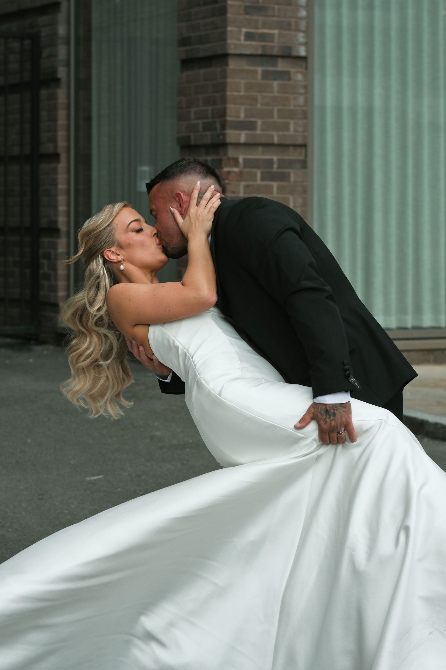Groom dipping and kissing bride in white gown on urban street after wedding.