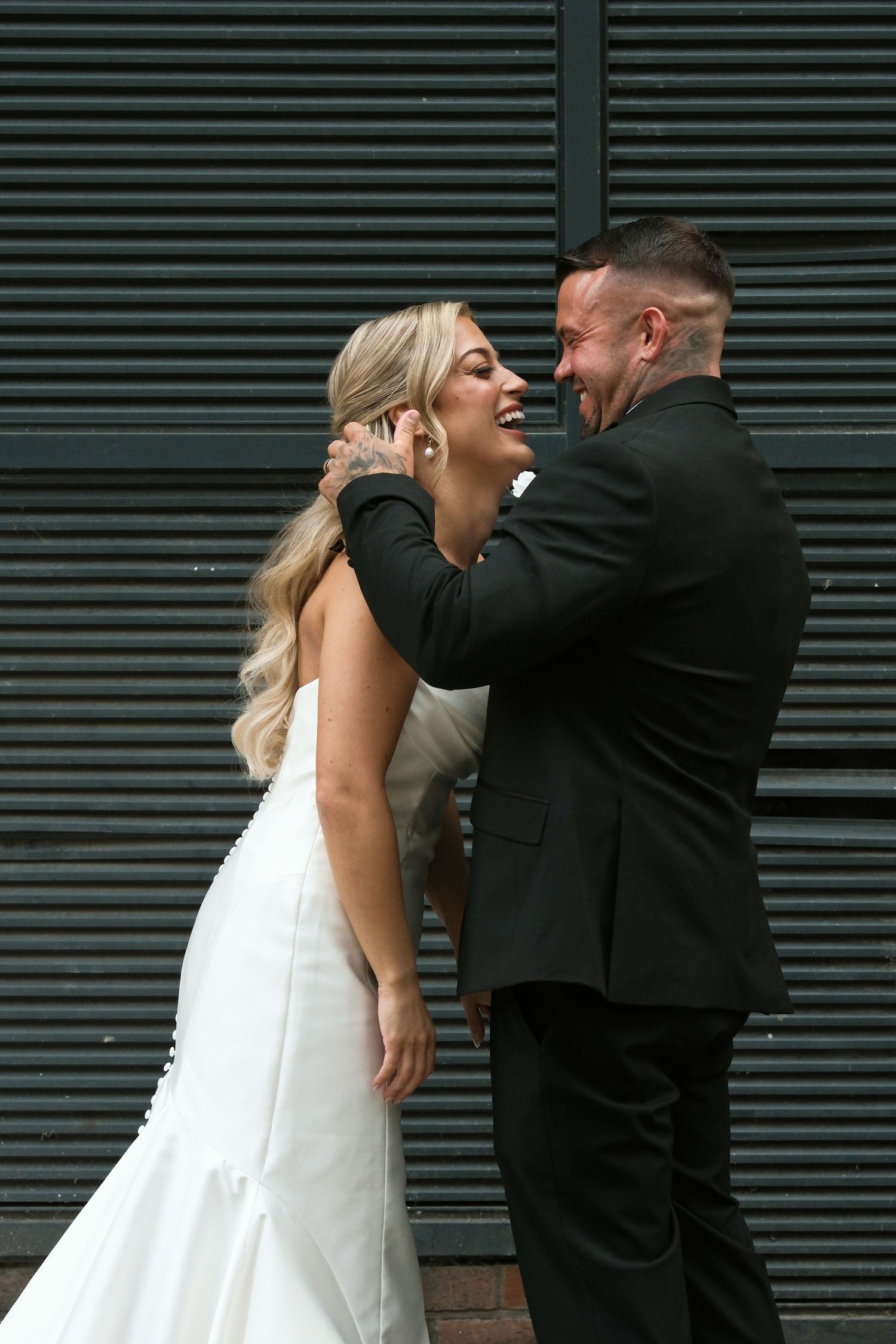 Laughing bride and groom embracing playfully in front of dark corrugated metal wall on wedding day.