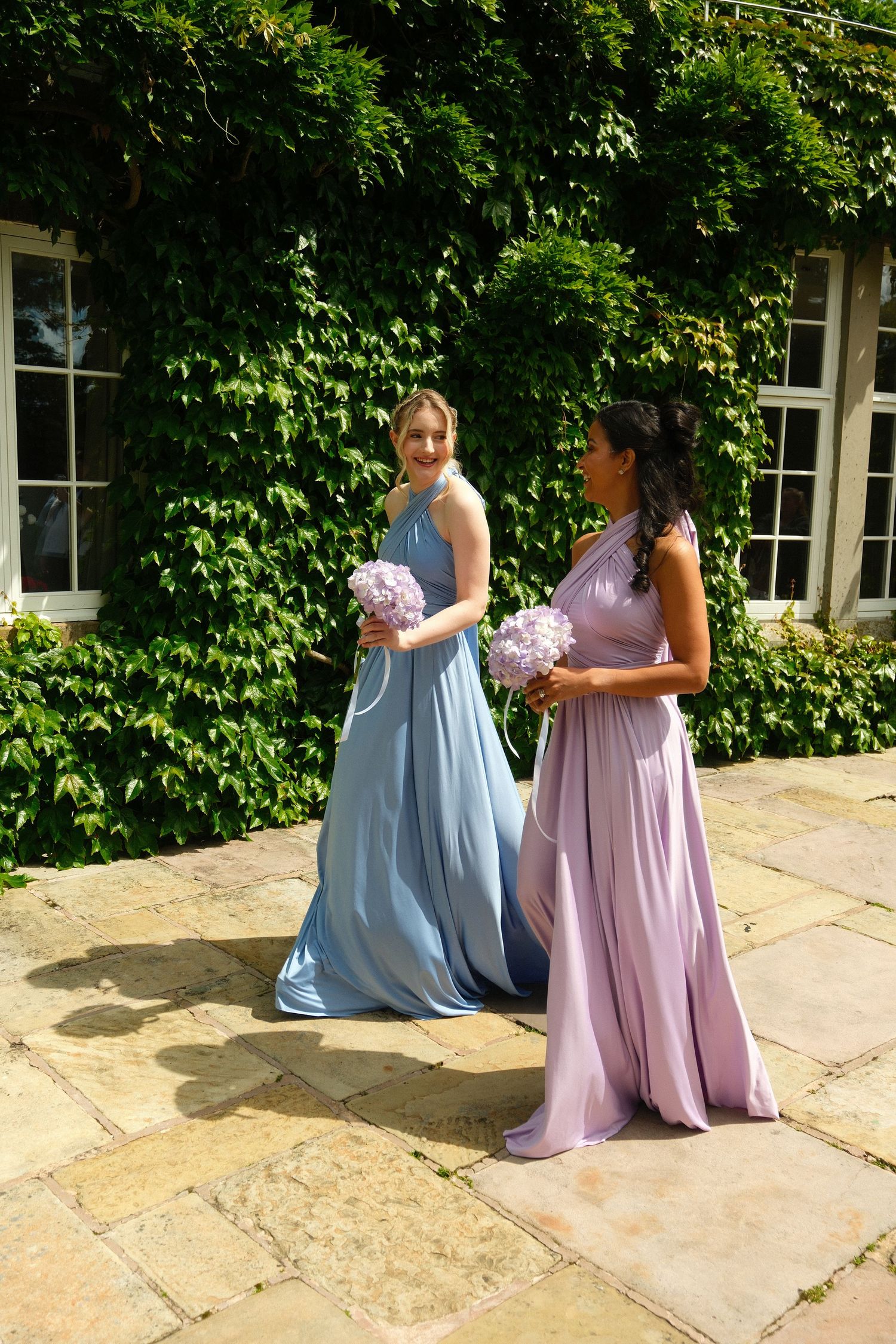 Two bridesmaids in blue and lavender gowns holding pink hydrangea bouquets outside ivy-covered building.