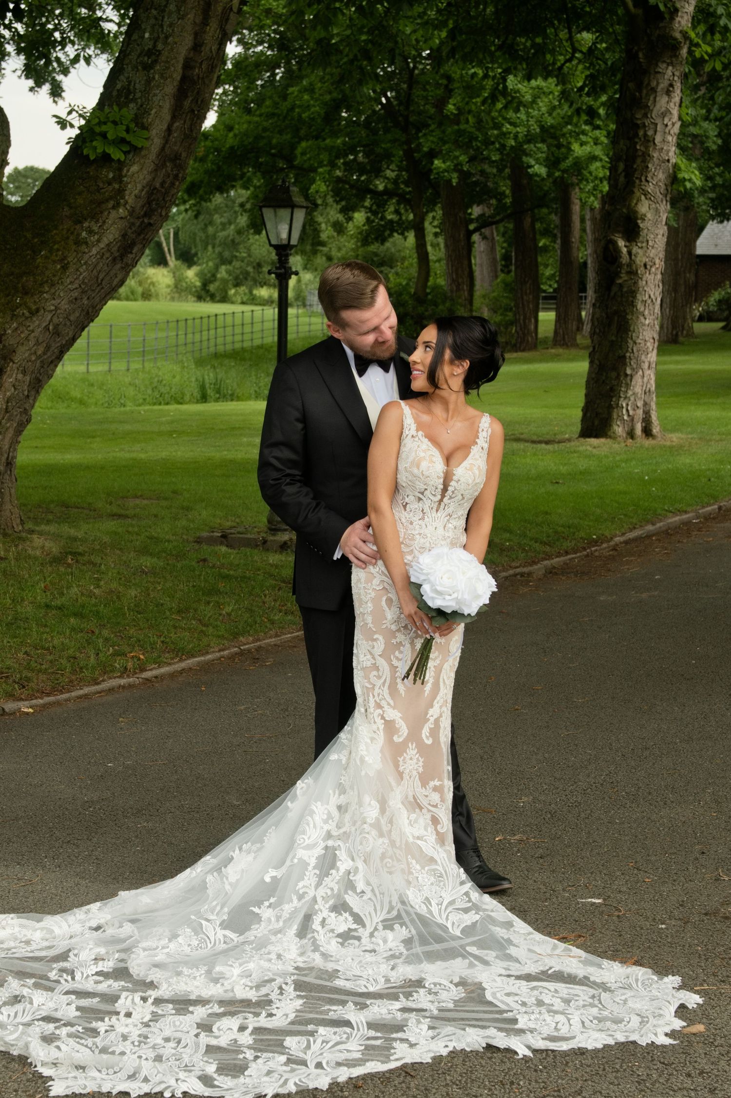 Bride in lace gown with train and groom in tuxedo embracing under trees.