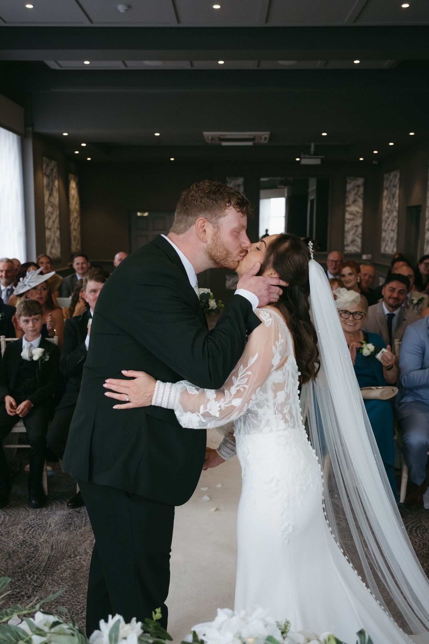 Bride and groom share their first kiss at the altar during an indoor wedding ceremony with guests seated behind.