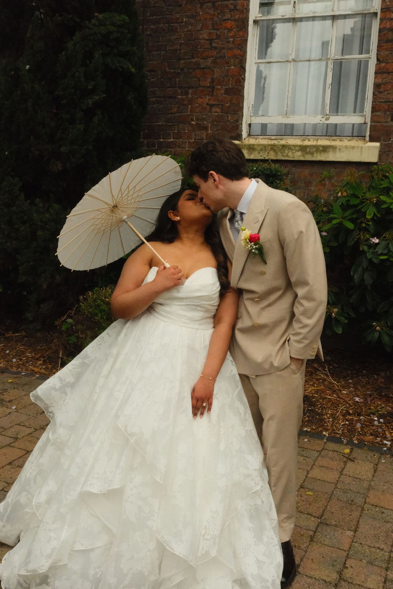 Groom kisses bride on cheek as she holds a white parasol, standing together in front of brick building.