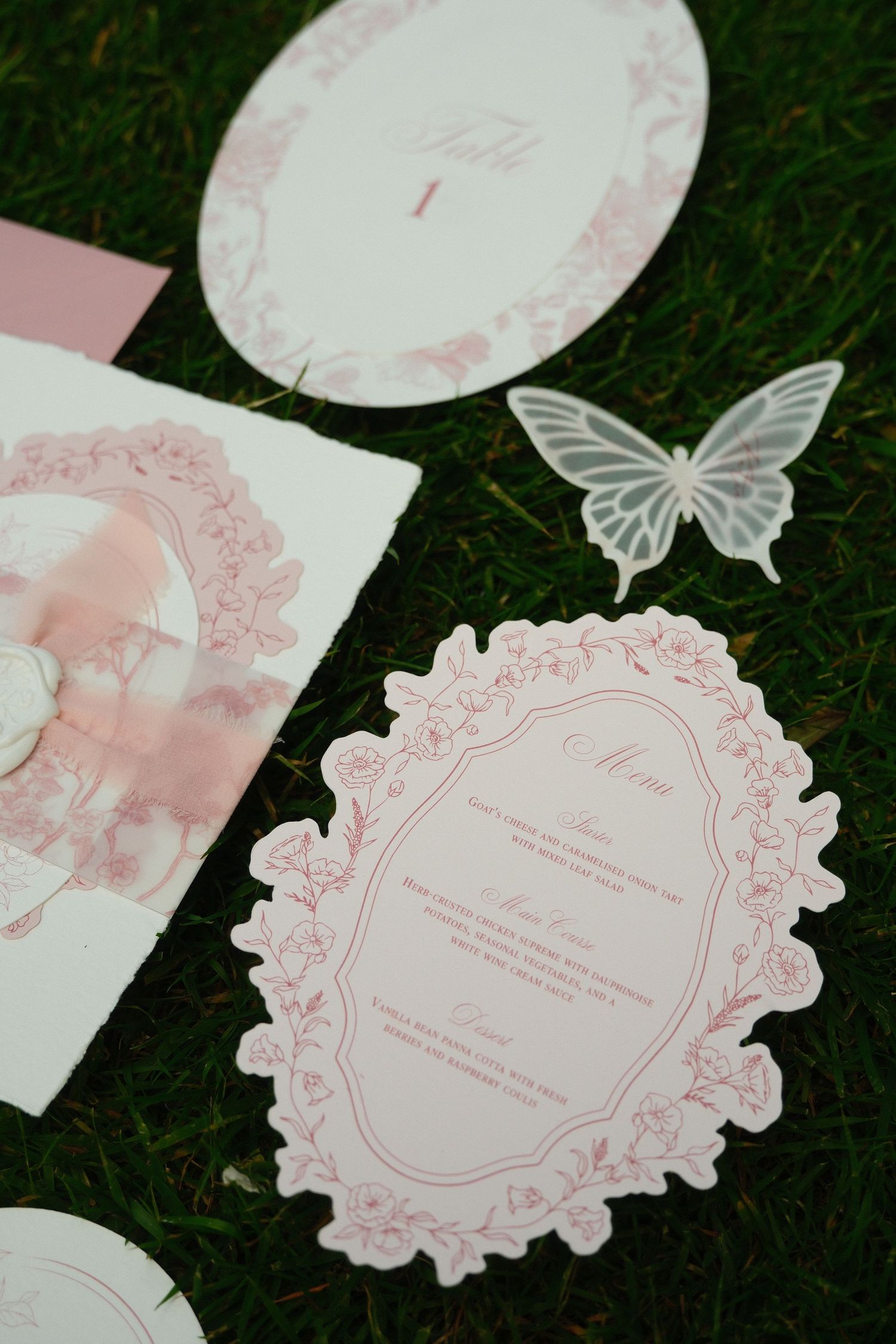 Pink floral wedding stationery suite with menu cards and butterfly accents displayed on green grass.
