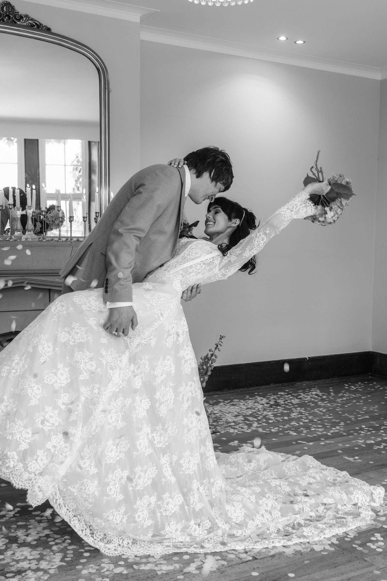 Black and white romantic photo of groom dipping bride in lace wedding dress during indoor celebration.