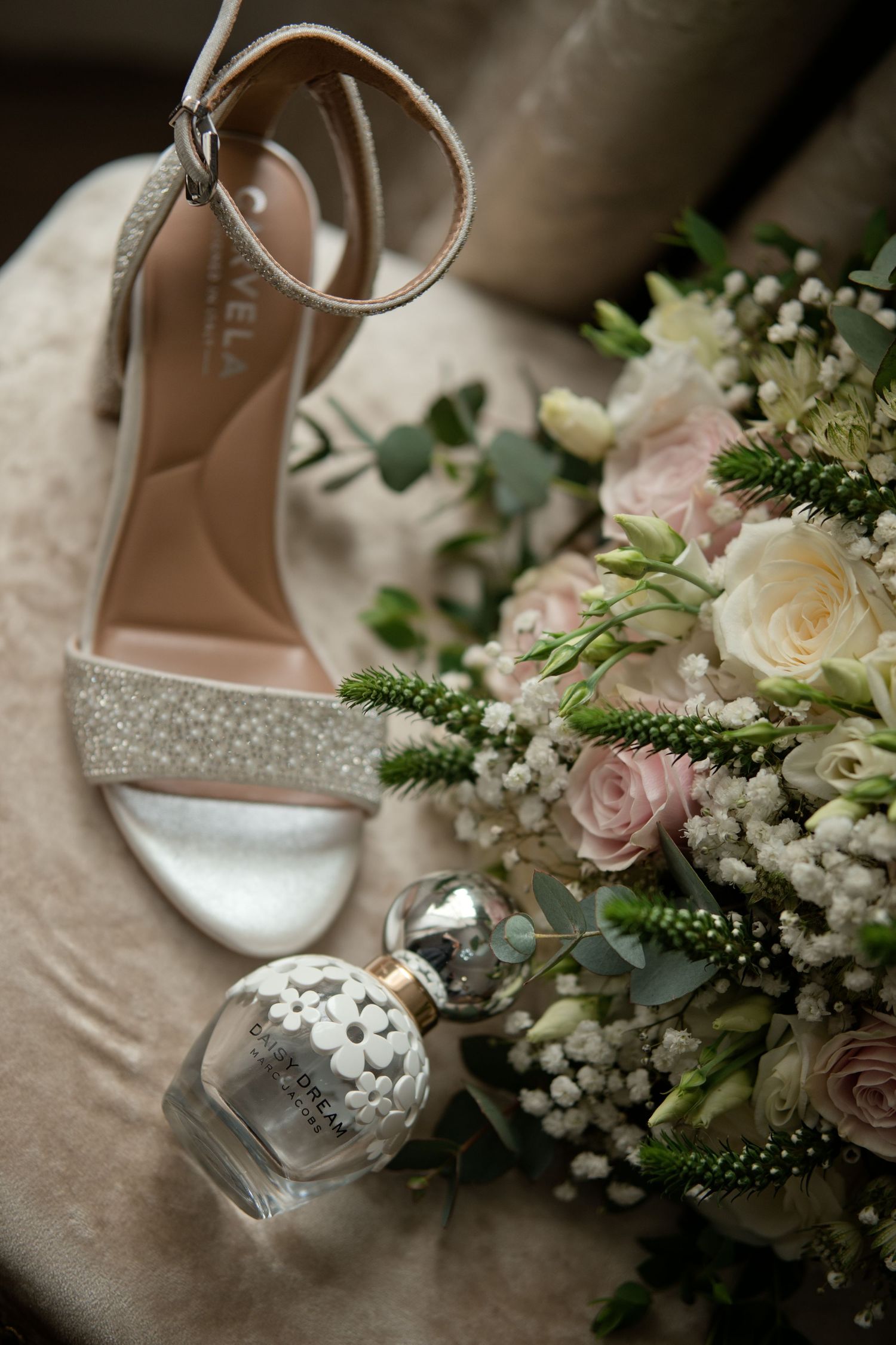 Elegant bridal heels, perfume bottle, and pink and white wedding bouquet on velvet chair.