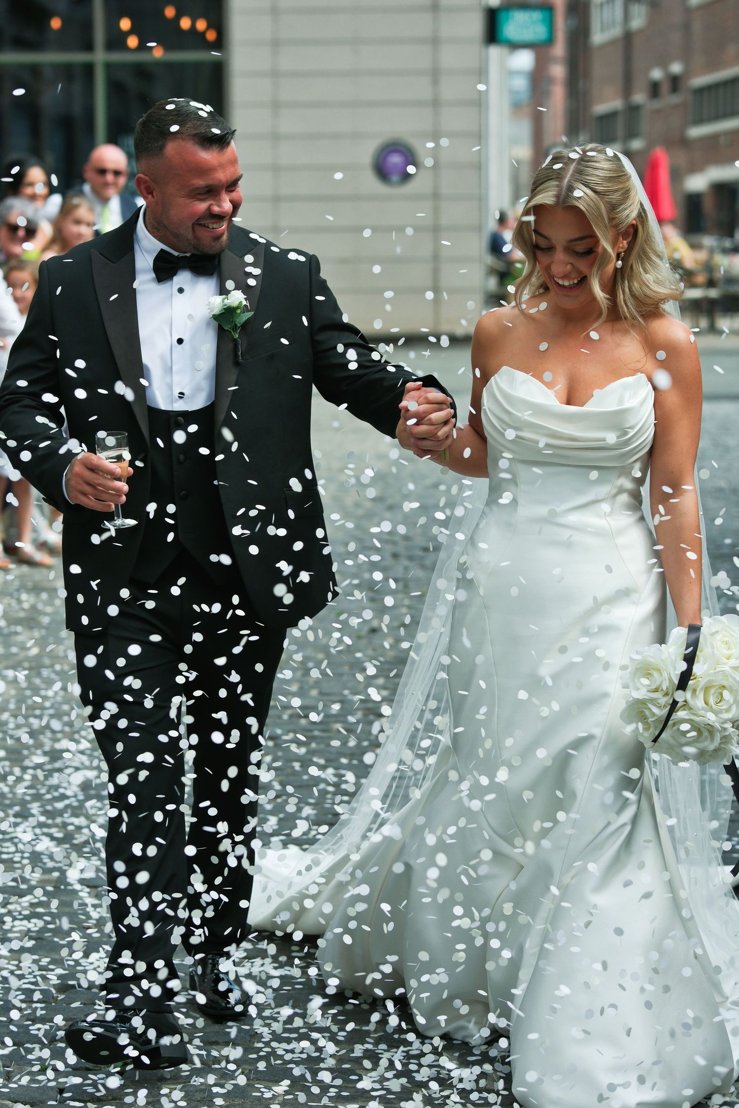 Smiling newlyweds holding hands walking through white confetti shower on urban street.