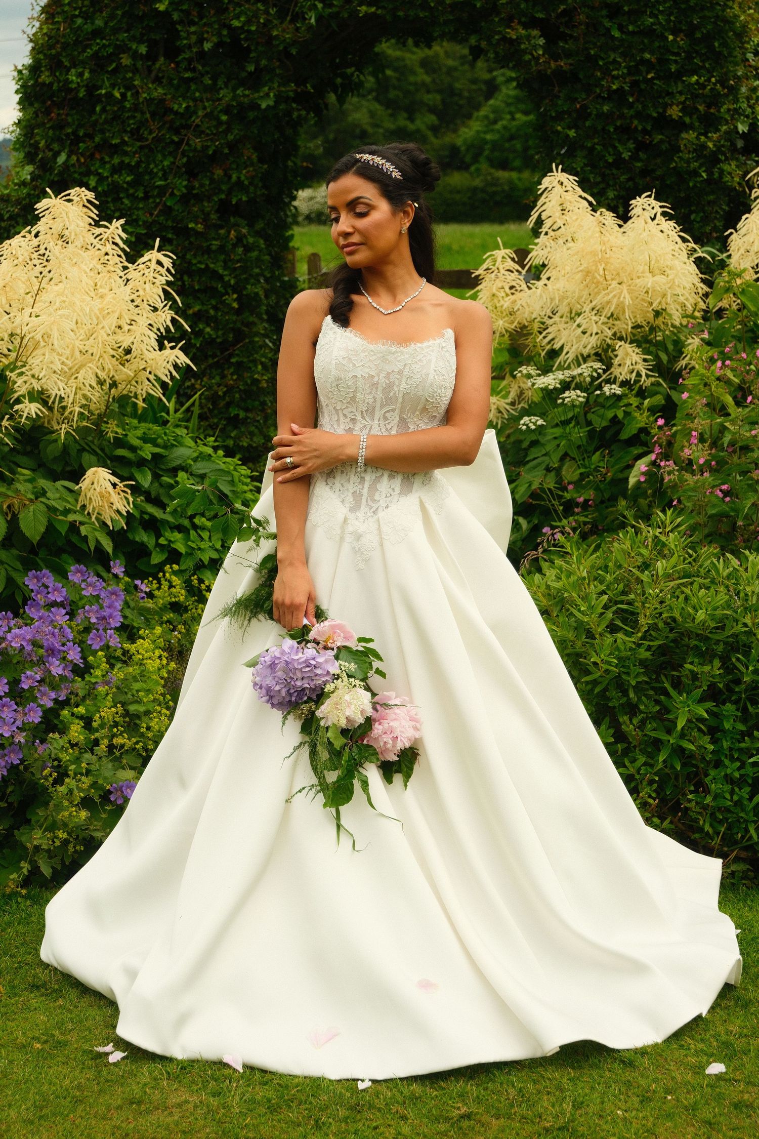 Beautiful bride in strapless white ball gown holding pink and purple hydrangea bouquet in lush garden setting.