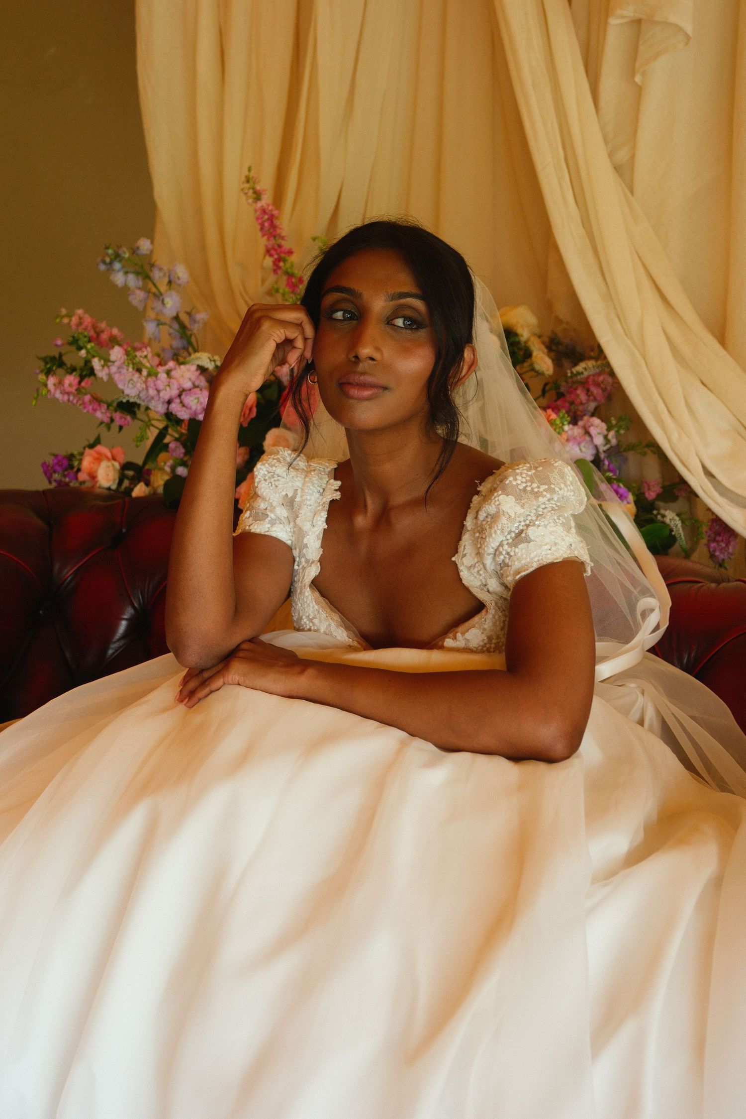 Elegant bride in white ballgown sits thoughtfully on sofa surrounded by floral decorations.