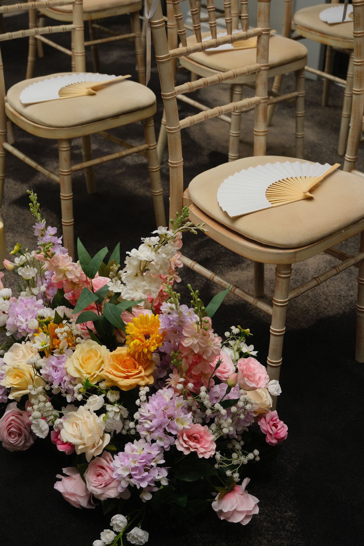 Wedding ceremony chairs with hand fans beside colorful floral arrangement on dark floor.