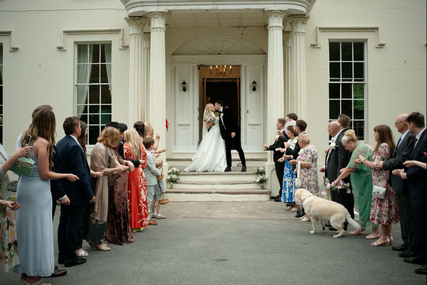 Wedding couple kissing on steps of white manor house with guests lining path.