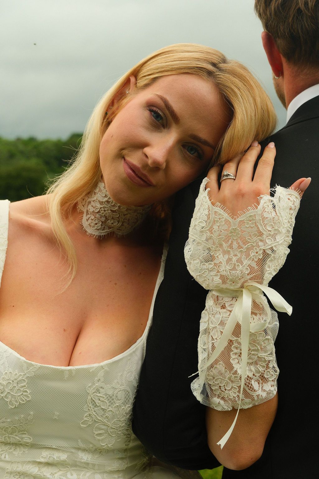 Blonde bride in white lace dress and gloves leans on groom's shoulder, smiling softly outdoors.