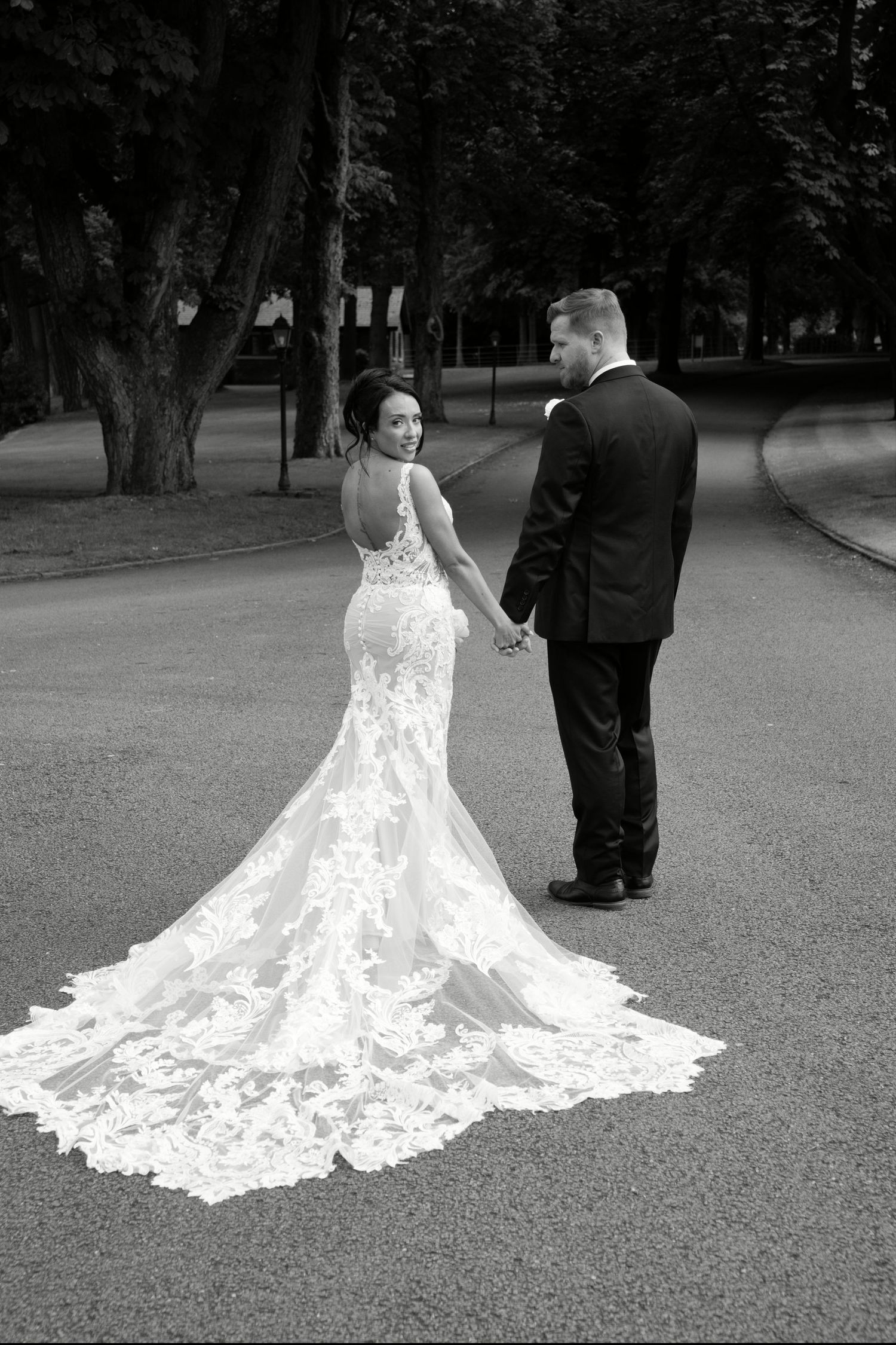 Bride in backless lace mermaid gown with long train holds grooms hand on a tree-lined path.