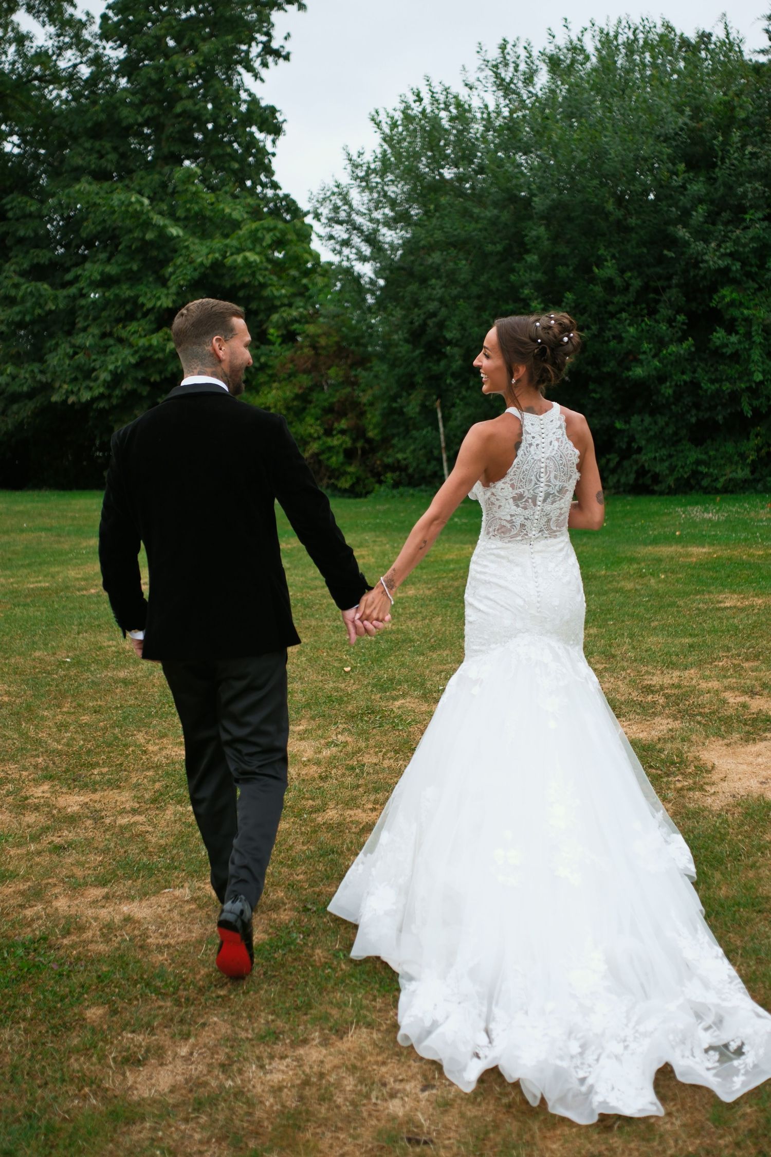 Bride and groom walking hand in hand across green lawn, bride in lace mermaid gown, groom in black suit with red shoes.