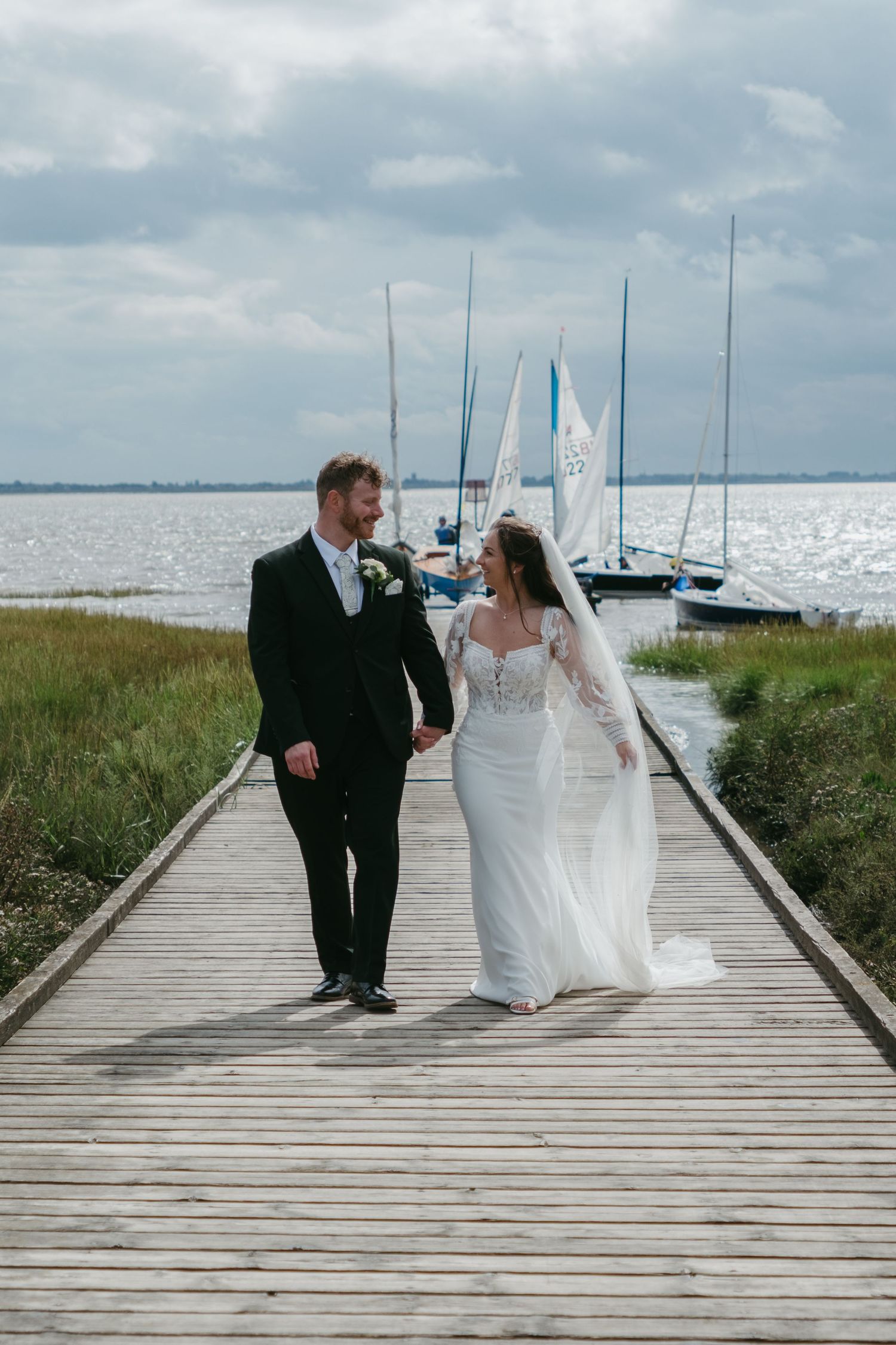 Bride and groom walk hand-in-hand on a wooden pier with sailboats and water in the background on their wedding day.