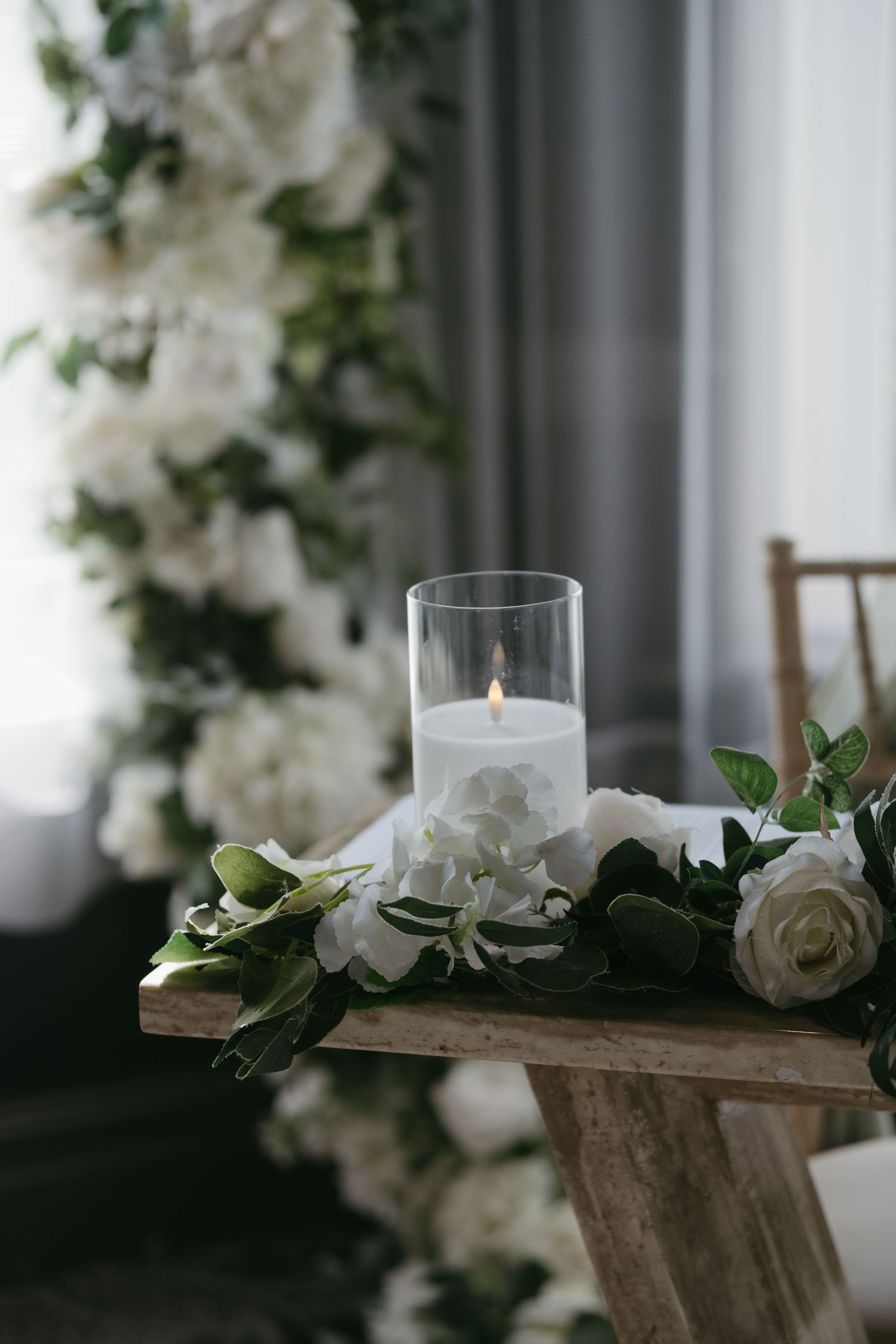 Elegant wedding candle in glass holder surrounded by white roses and greenery on a rustic wooden bench.