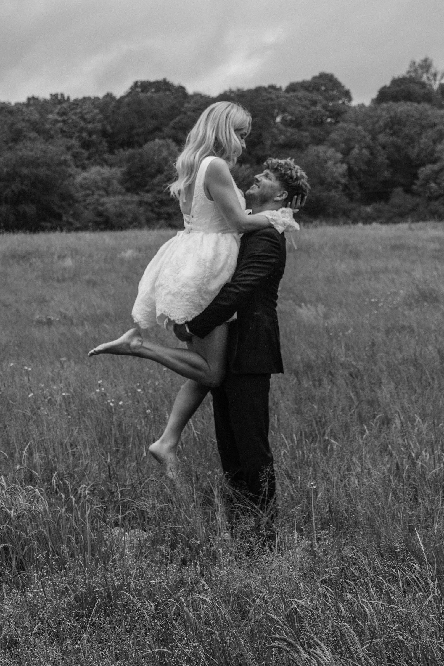 Groom lifting bride in white dress in a grassy field, romantic black and white wedding photo.