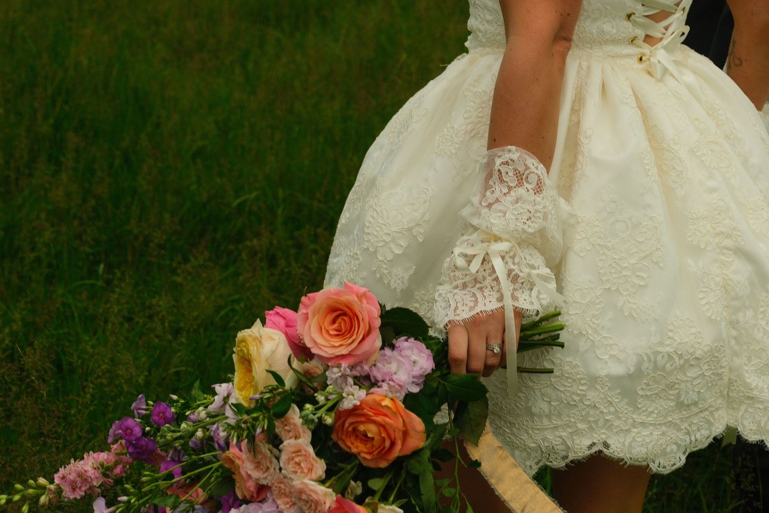 Bride in short lace wedding dress holds colorful bouquet of roses and wildflowers.