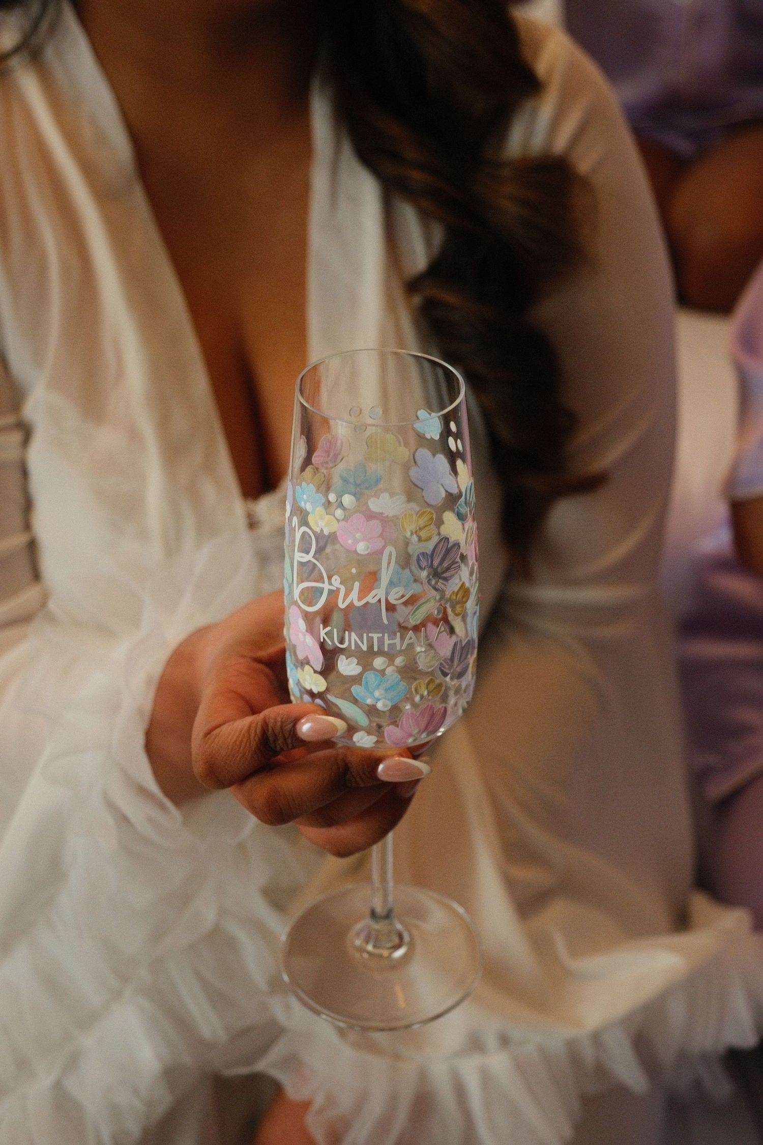 Bride holds personalized floral champagne glass labeled Bride Kuntha at wedding celebration.