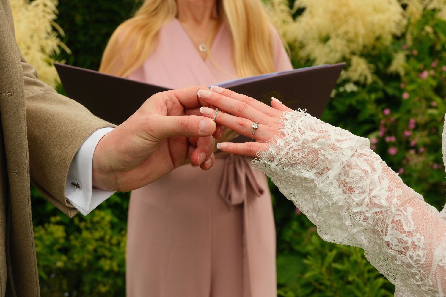 Bride and groom exchanging rings during wedding ceremony, officiant holding book in background.