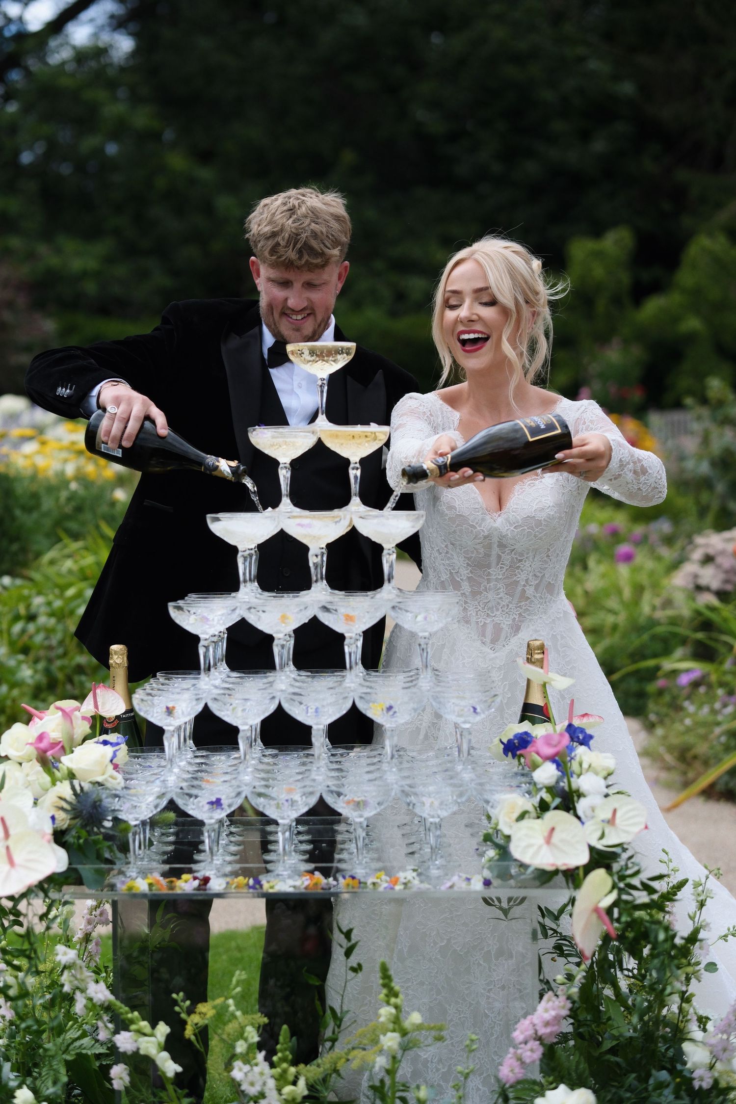 Bride and groom pouring champagne into a elegant glass tower surrounded by flowers at their wedding reception.