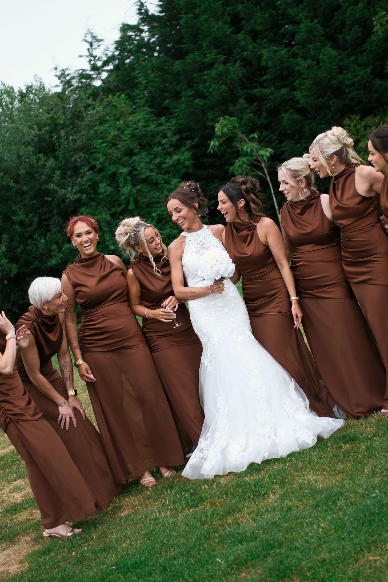 Bride in white lace gown poses with bridesmaids in brown satin dresses on green lawn at outdoor wedding.