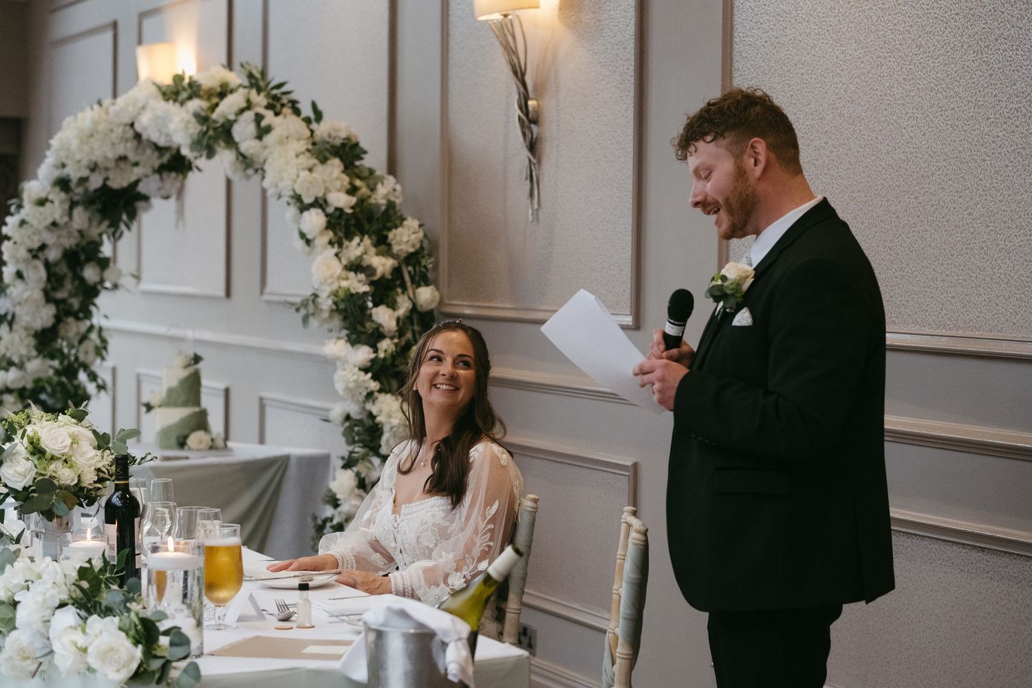 Groom gives wedding speech holding microphone beside floral arch while bride smiles at reception dinner table.