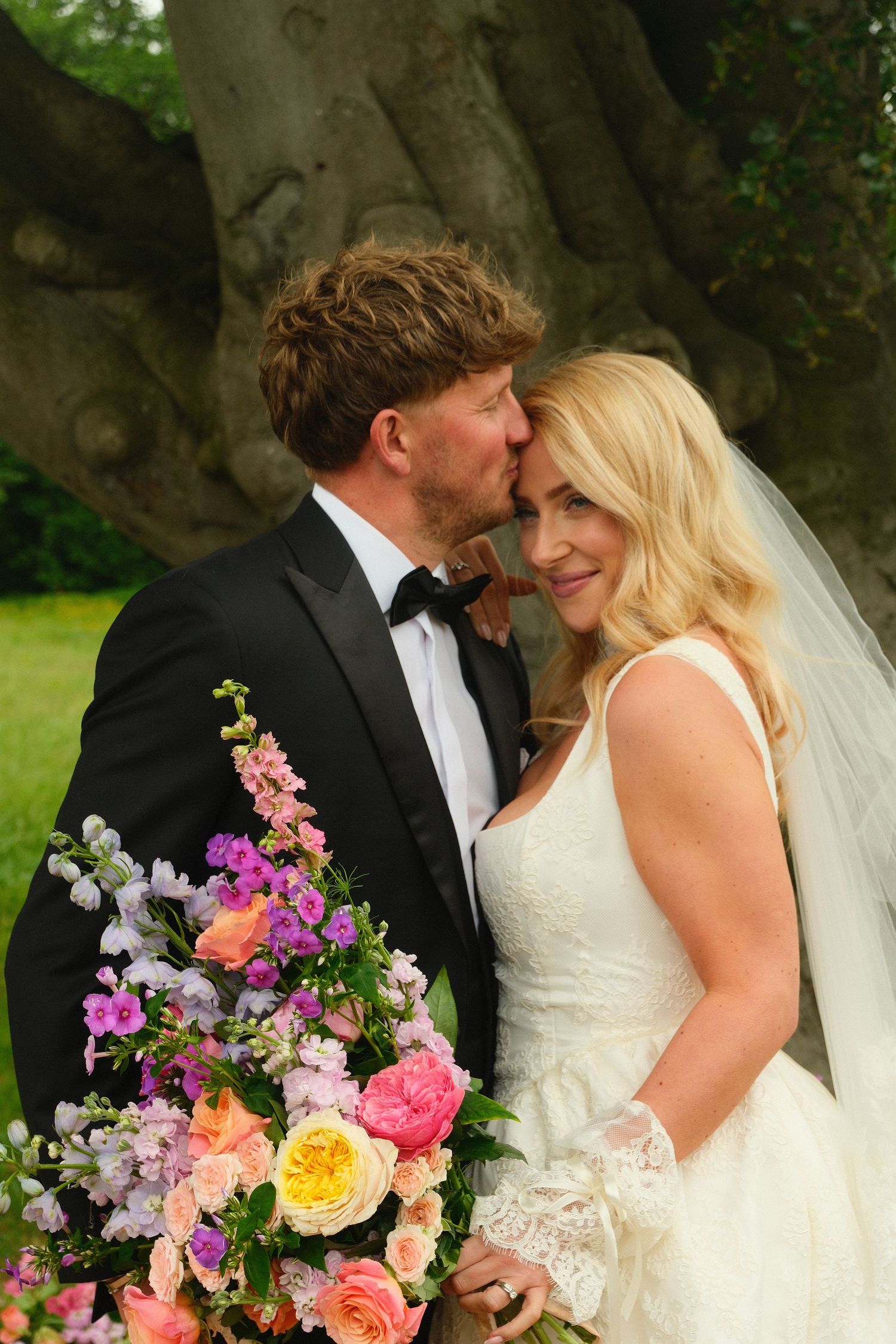 Groom in black tuxedo kissing blonde bride wearing lace wedding dress beside an ancient tree.