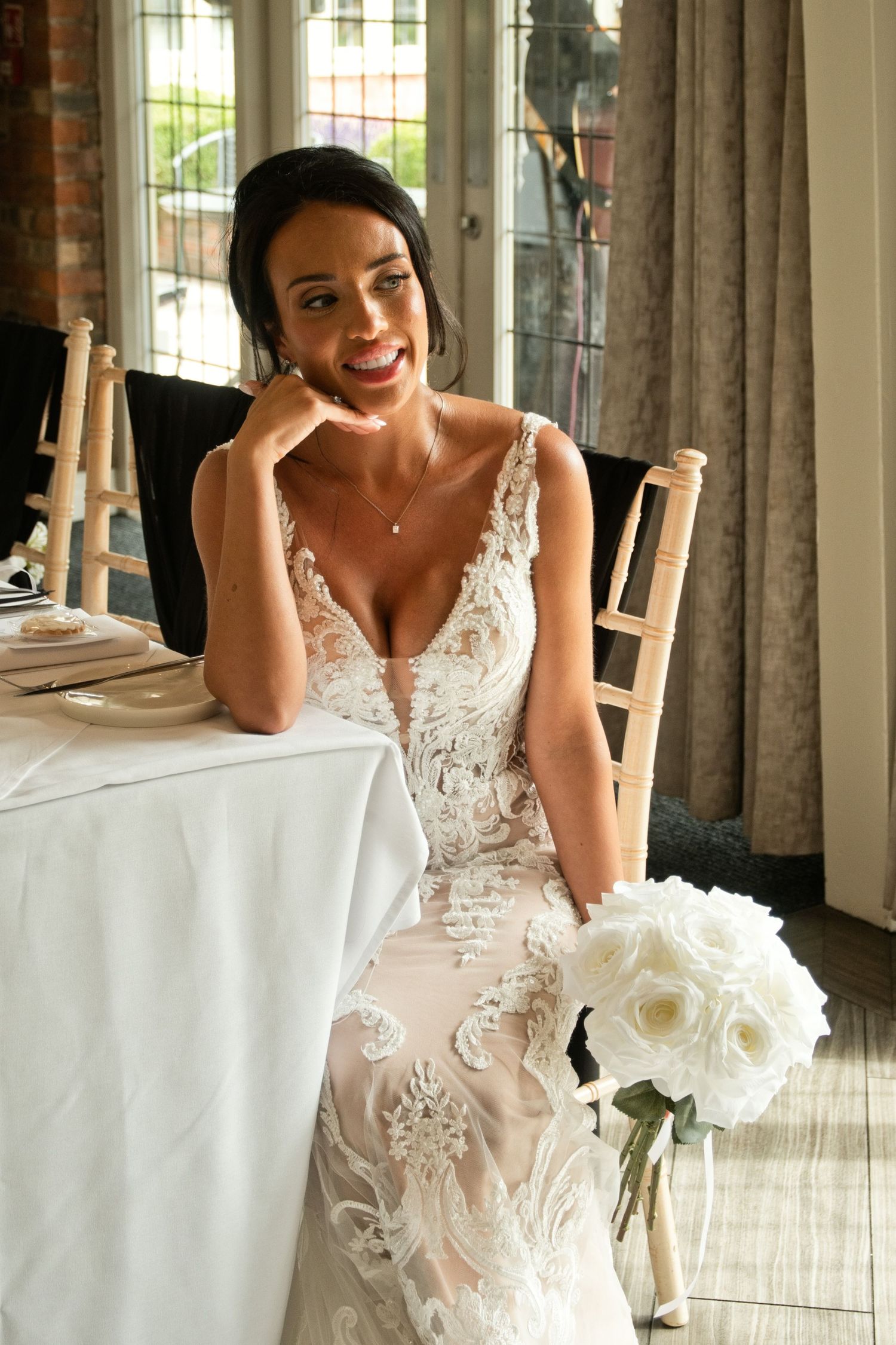 Beautiful dark-haired bride in plunging lace wedding gown seated at reception table with white bouquet.