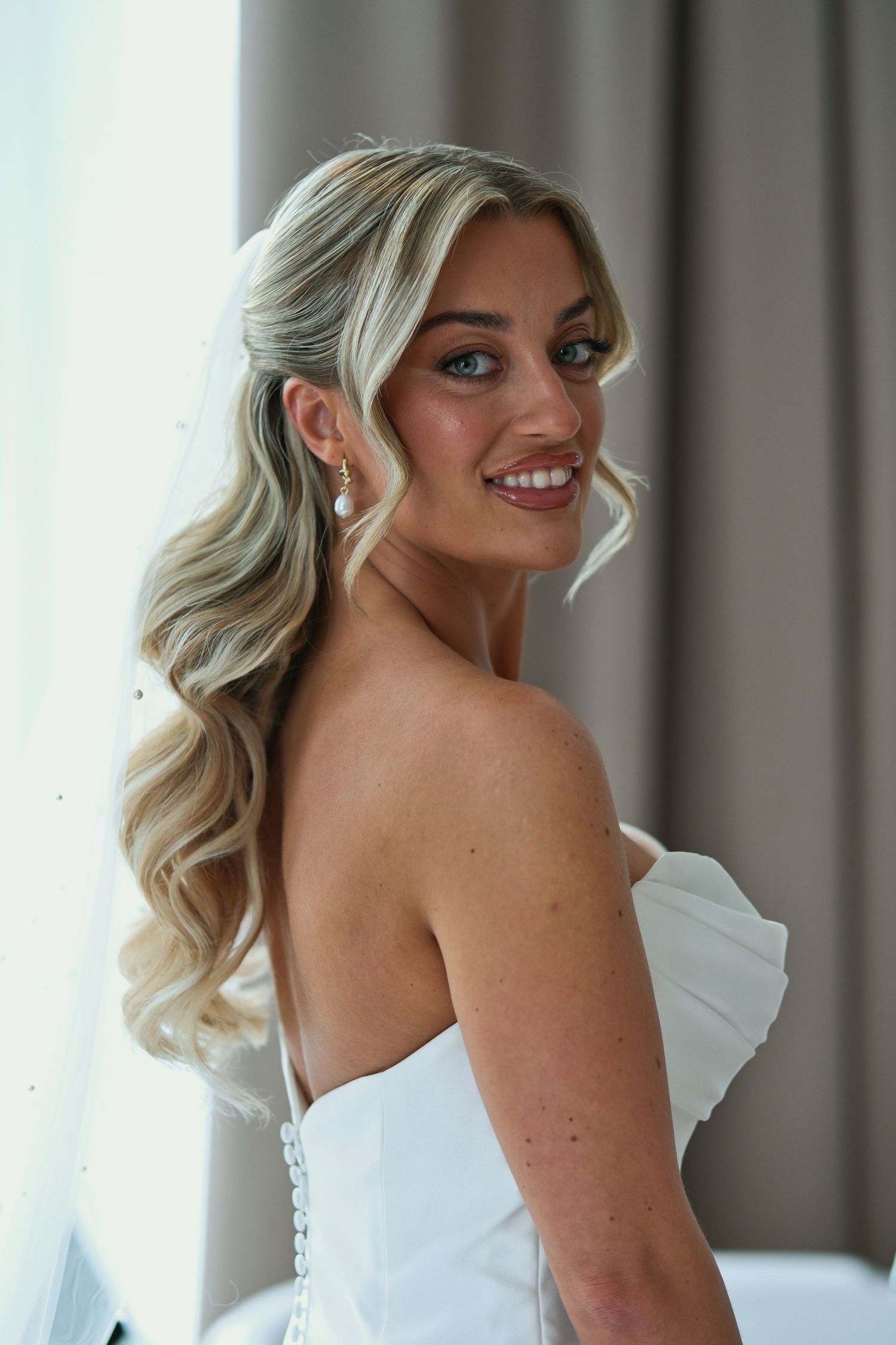 Radiant bride in strapless white gown with veil and wavy blonde hair smiling over her shoulder.