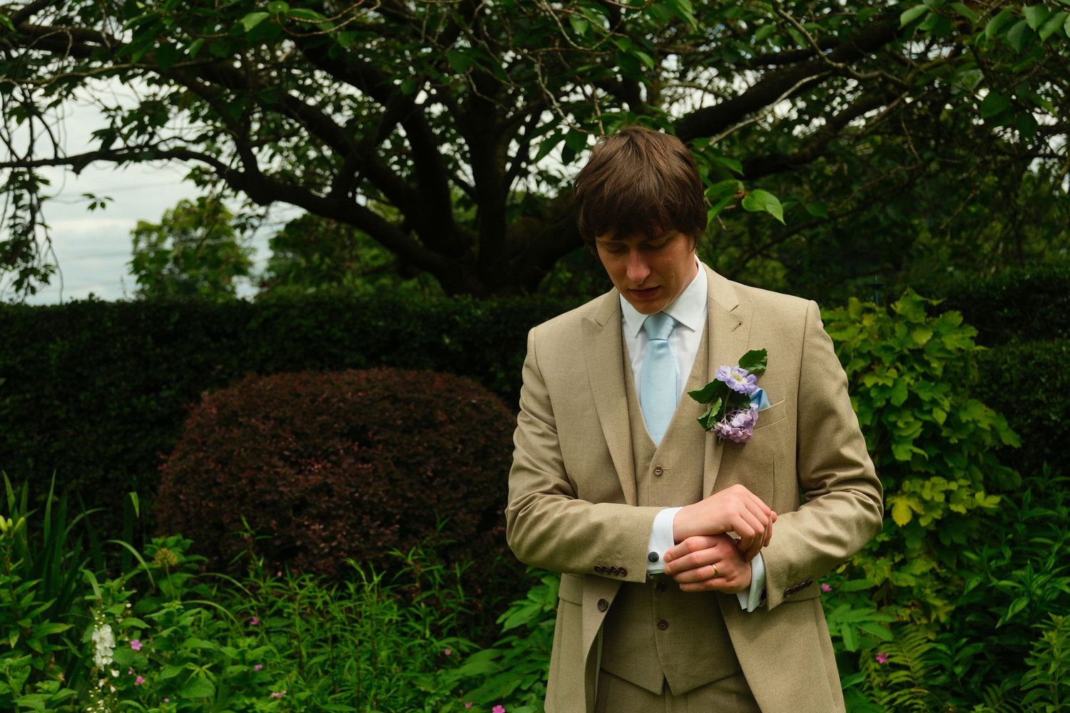 Groom in tan three-piece suit adjusts cufflinks in lush green garden before wedding ceremony.