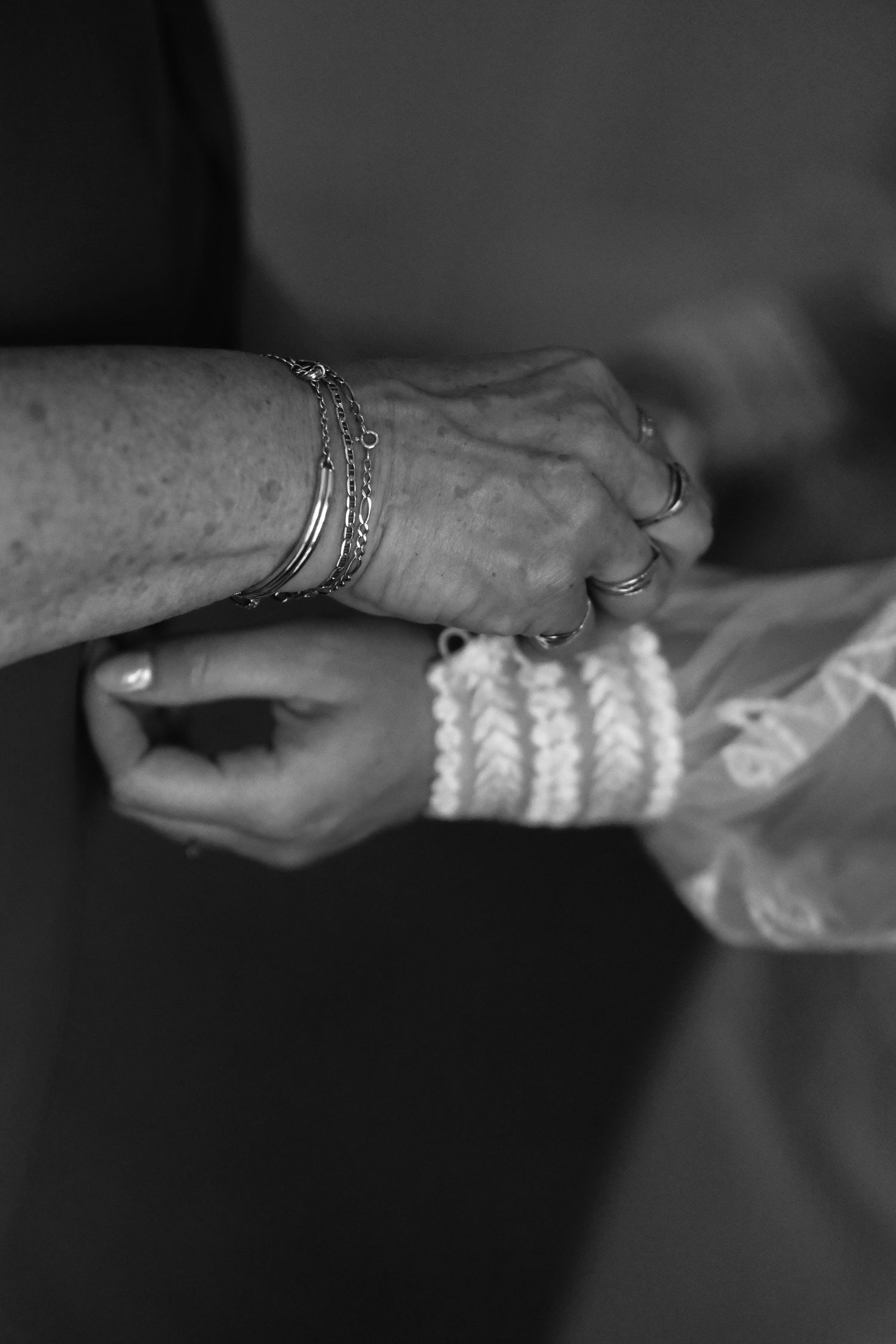 Black and white photo of hands fastening a braided bracelet on a wrist adorned with silver bangles and rings.