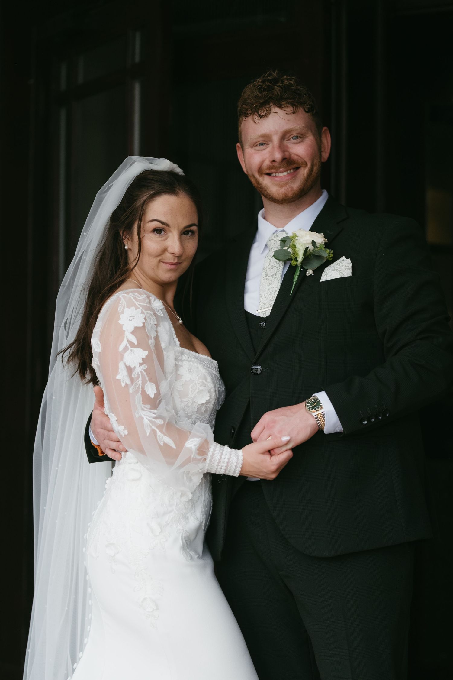 Smiling bride and groom posing together, bride in lace veil gown, groom in dark suit with boutonniere.