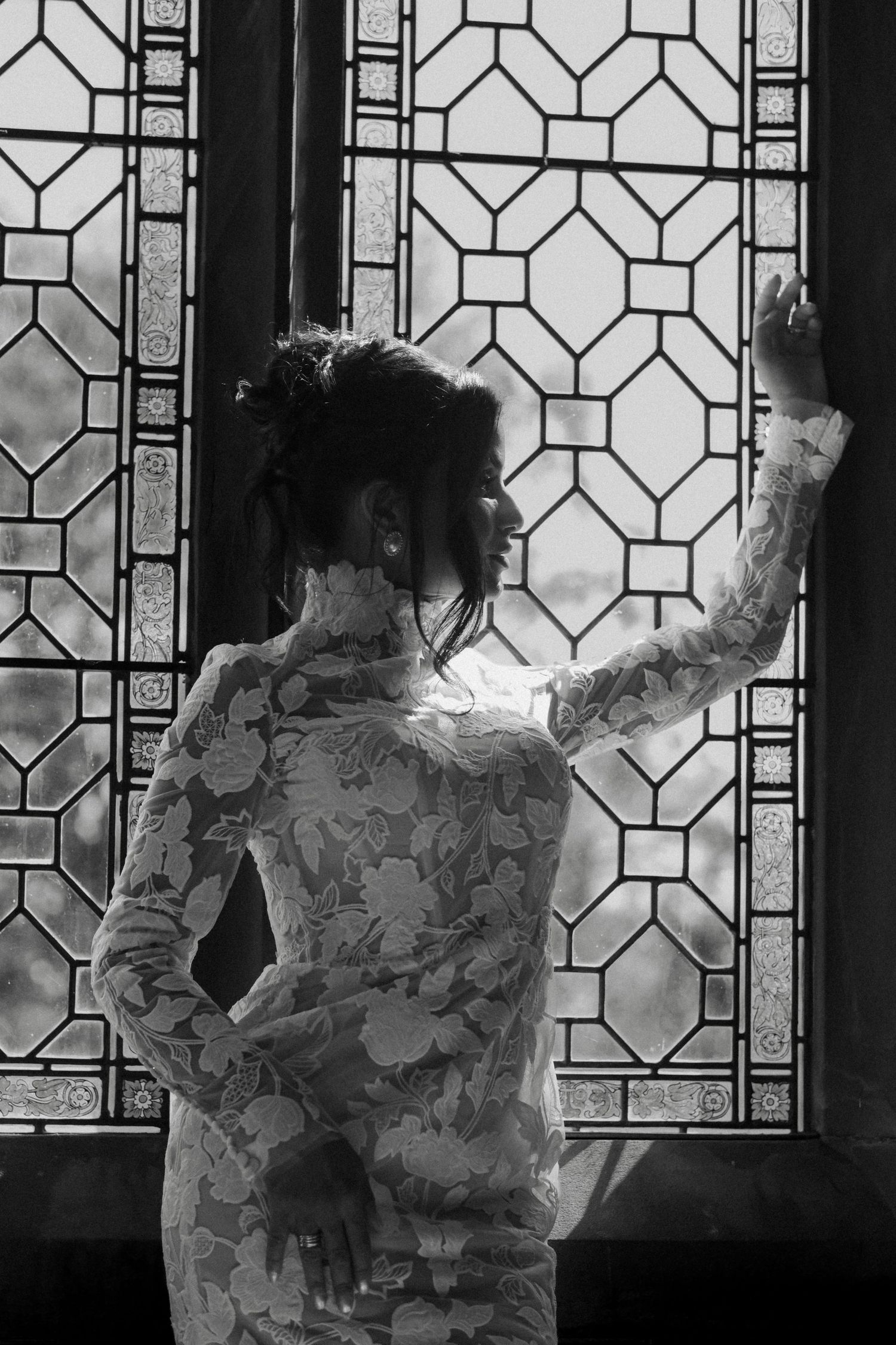Black and white photo of a bride in lace dress gazing through ornate stained glass window.