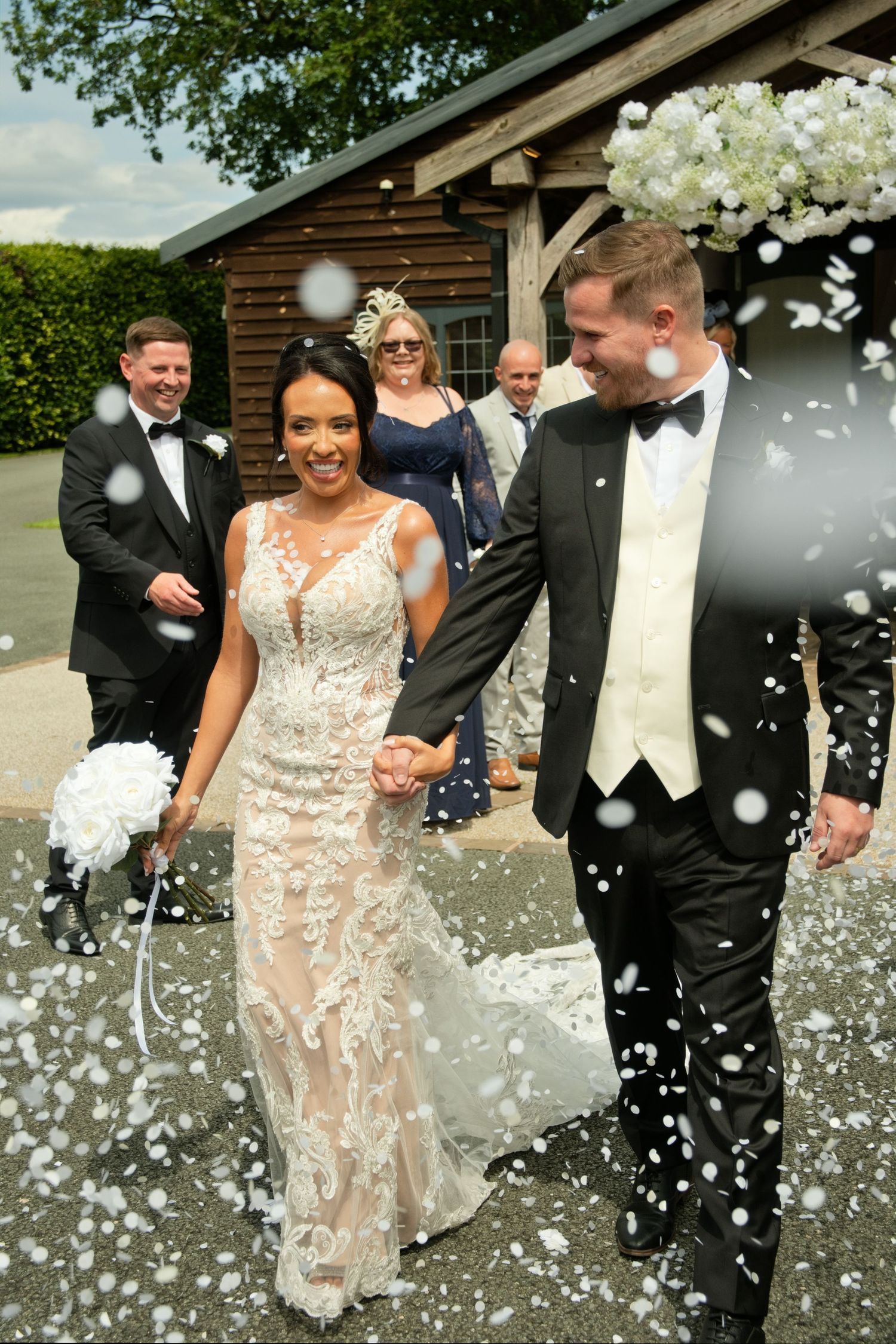 Joyful newlyweds walk through white confetti shower with wedding guests celebrating behind them outdoors.