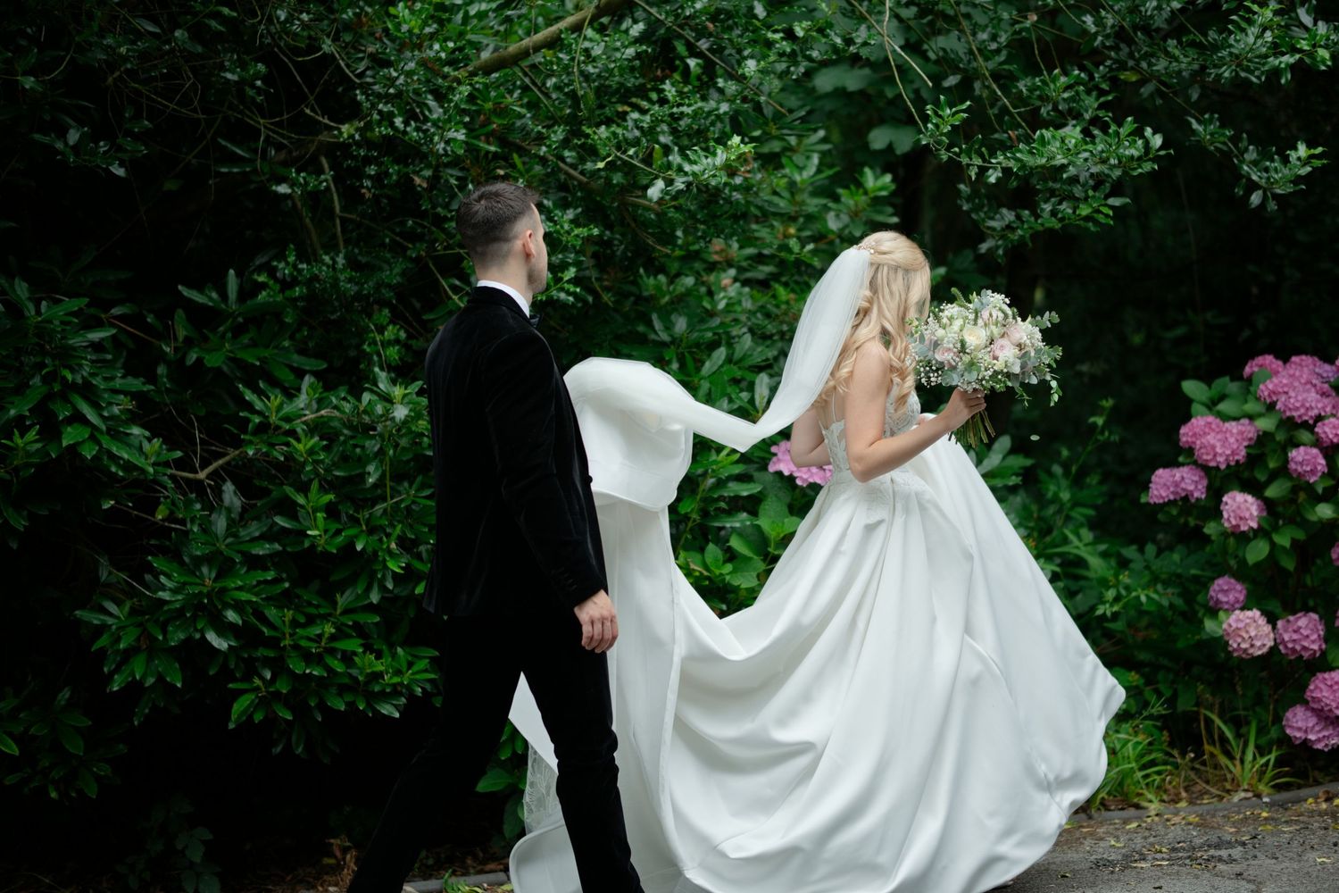 Bride and groom walking hand in hand through lush green garden, bride's white gown and veil flowing behind her.